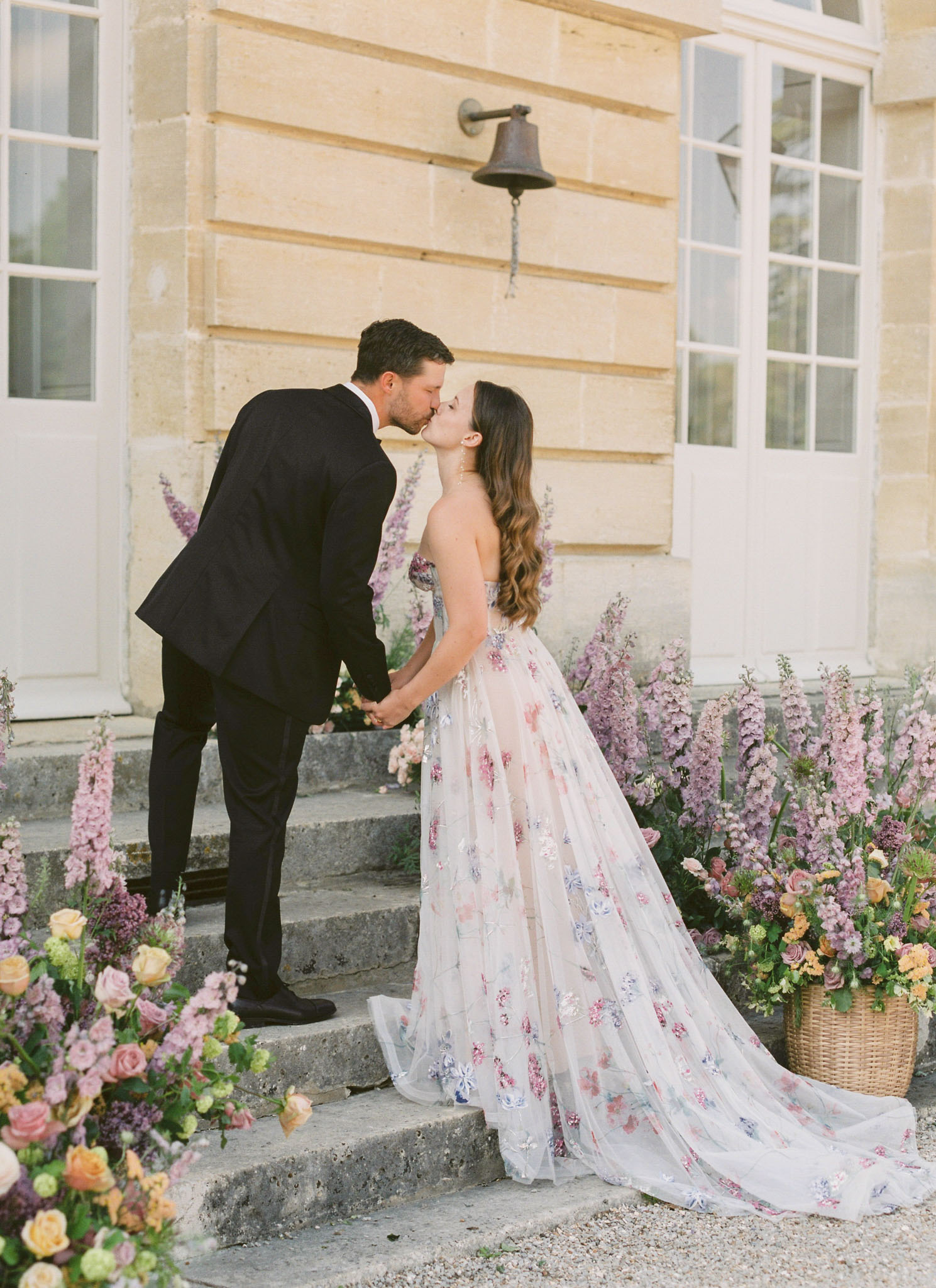 Bride and groom kissing on chateau steps surrounded by garden floral arrangements