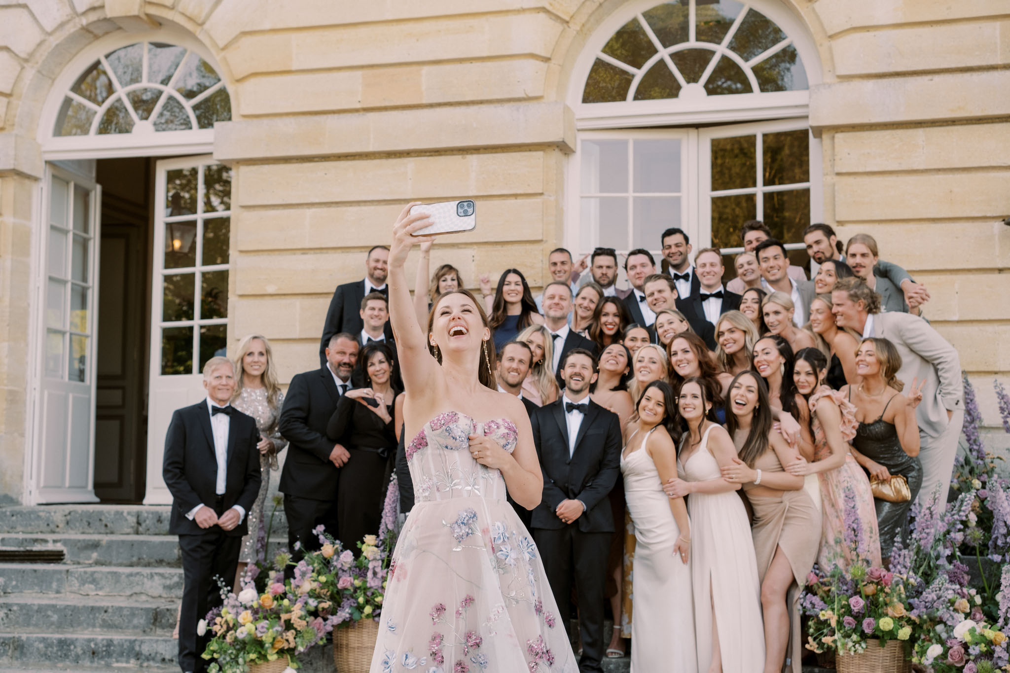 Bride taking group selfie with 40 guests on chateau stone steps flanked by pink and purple flower arrangements