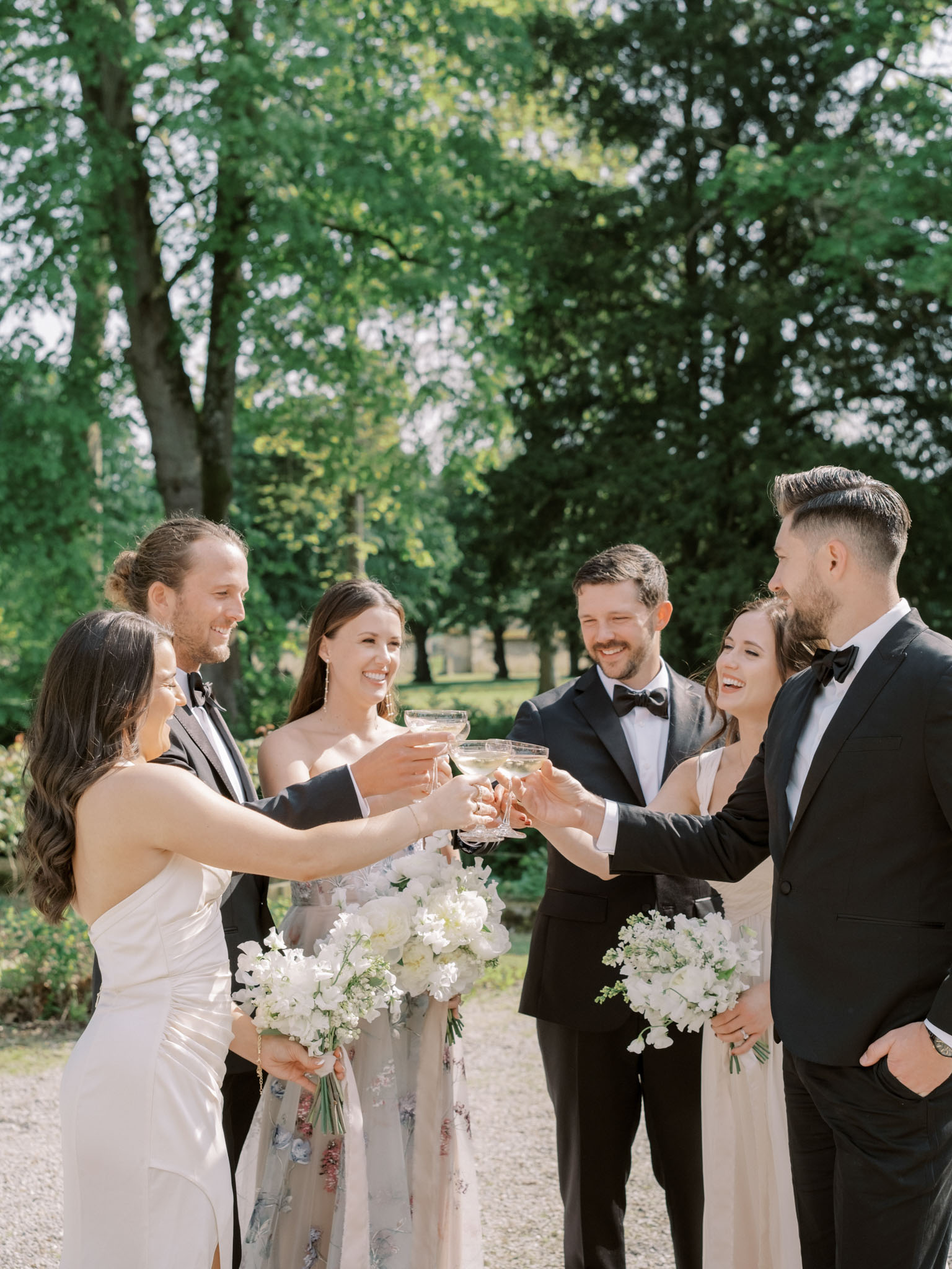 Three couples toasting with coupe glasses on gravel path during cocktail hour in garden setting