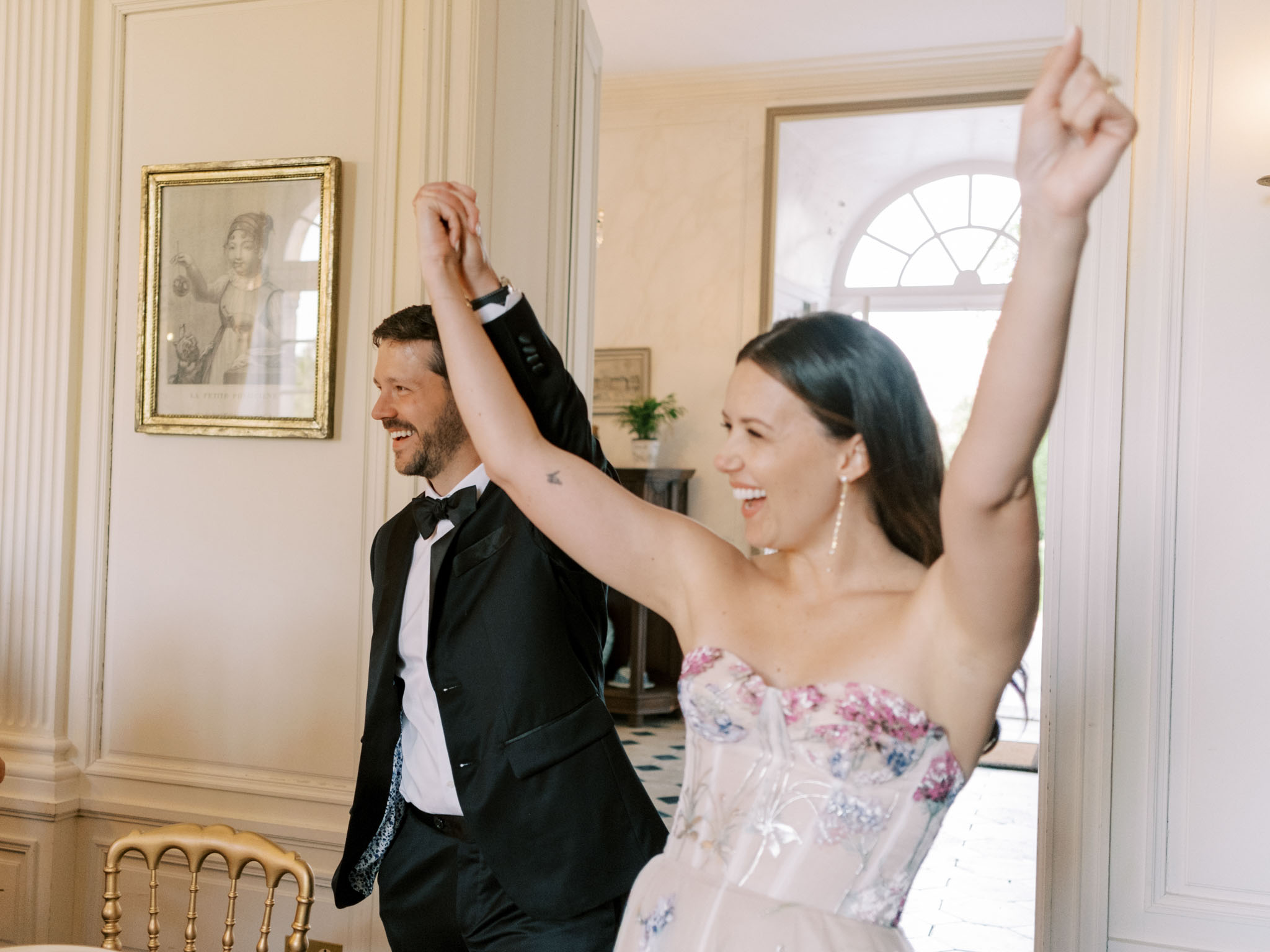 Couple making celebratory entrance into wood-panelled chateau reception room with arms raised
