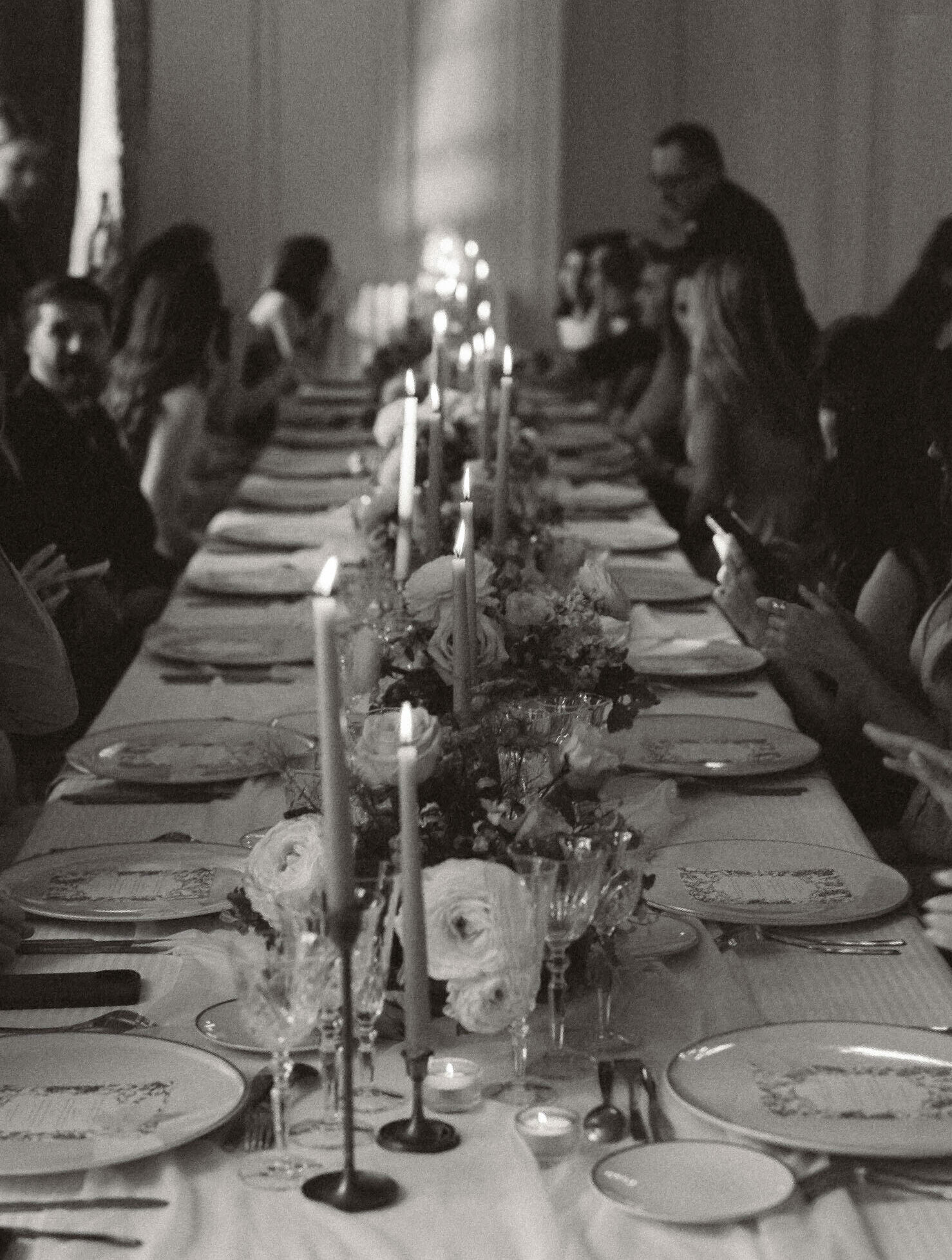 Black-and-white low-angle view of long banquet table with seated guests, taper candles, and floral centrepieces