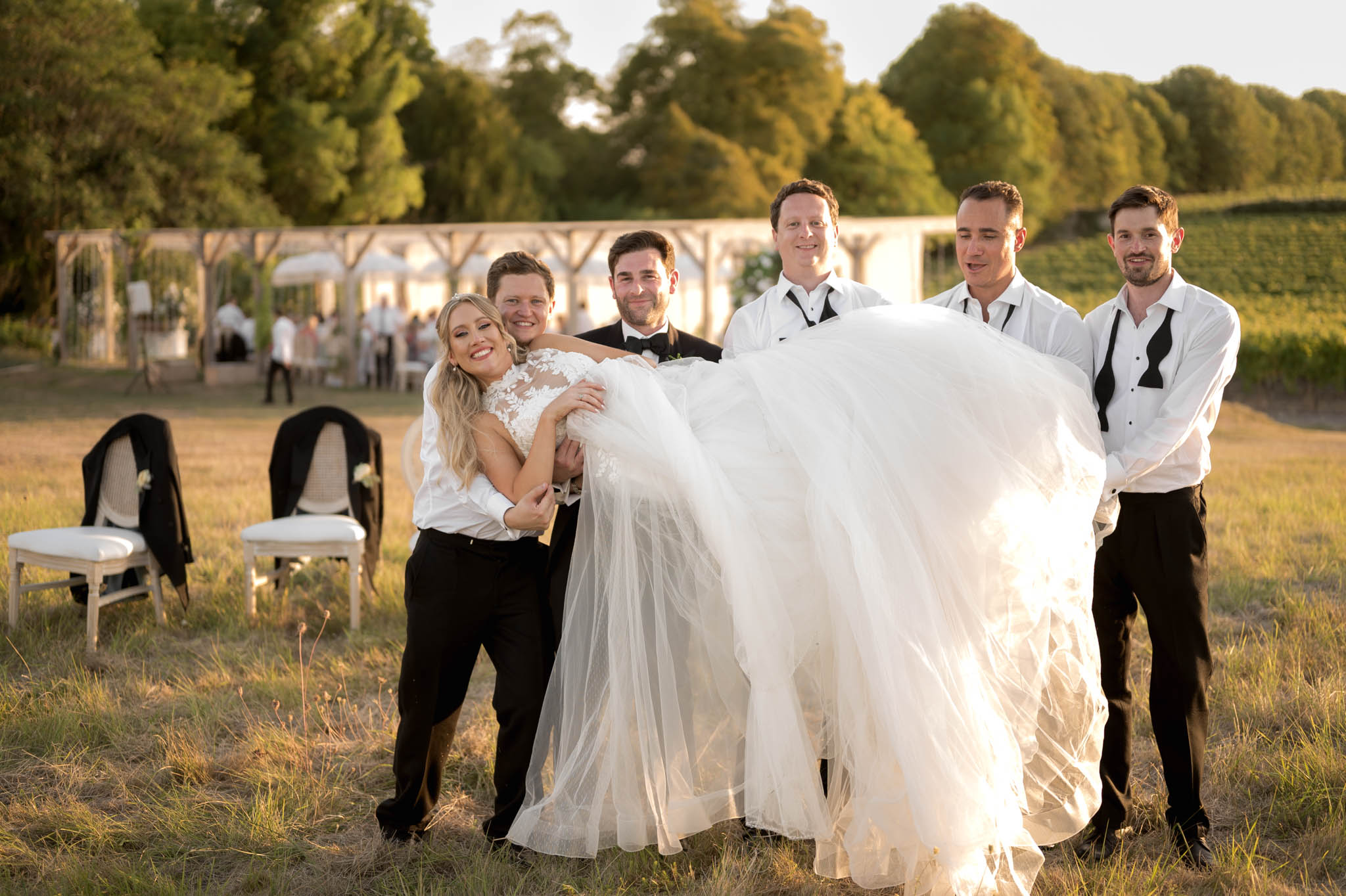 Groom and four groomsmen playfully fanning bride's tulle ball gown skirt at golden hour on estate grounds