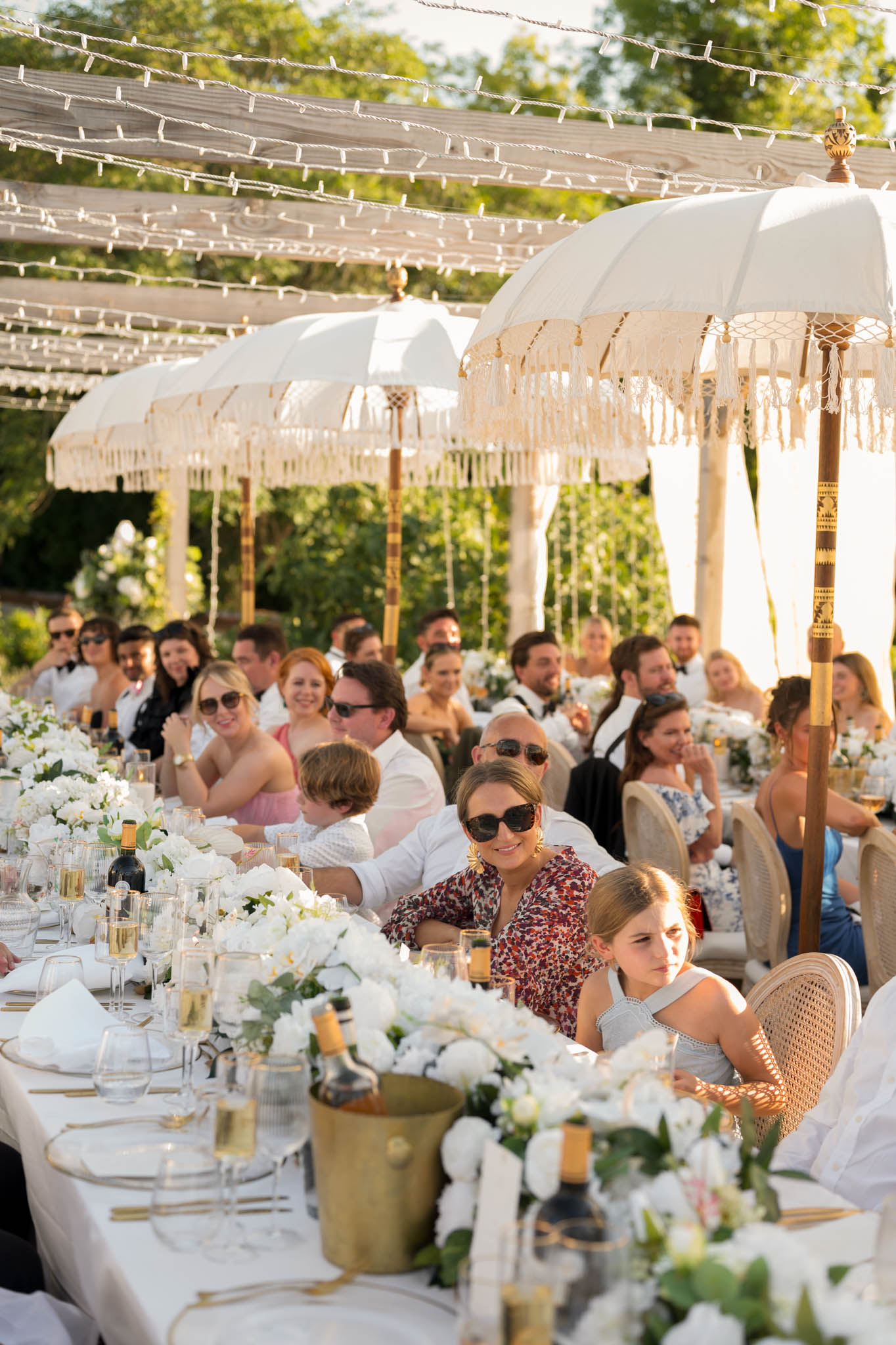 Outdoor banquet reception at golden hour with white rose centerpieces and fringed parasols under a fairy-lit pergola