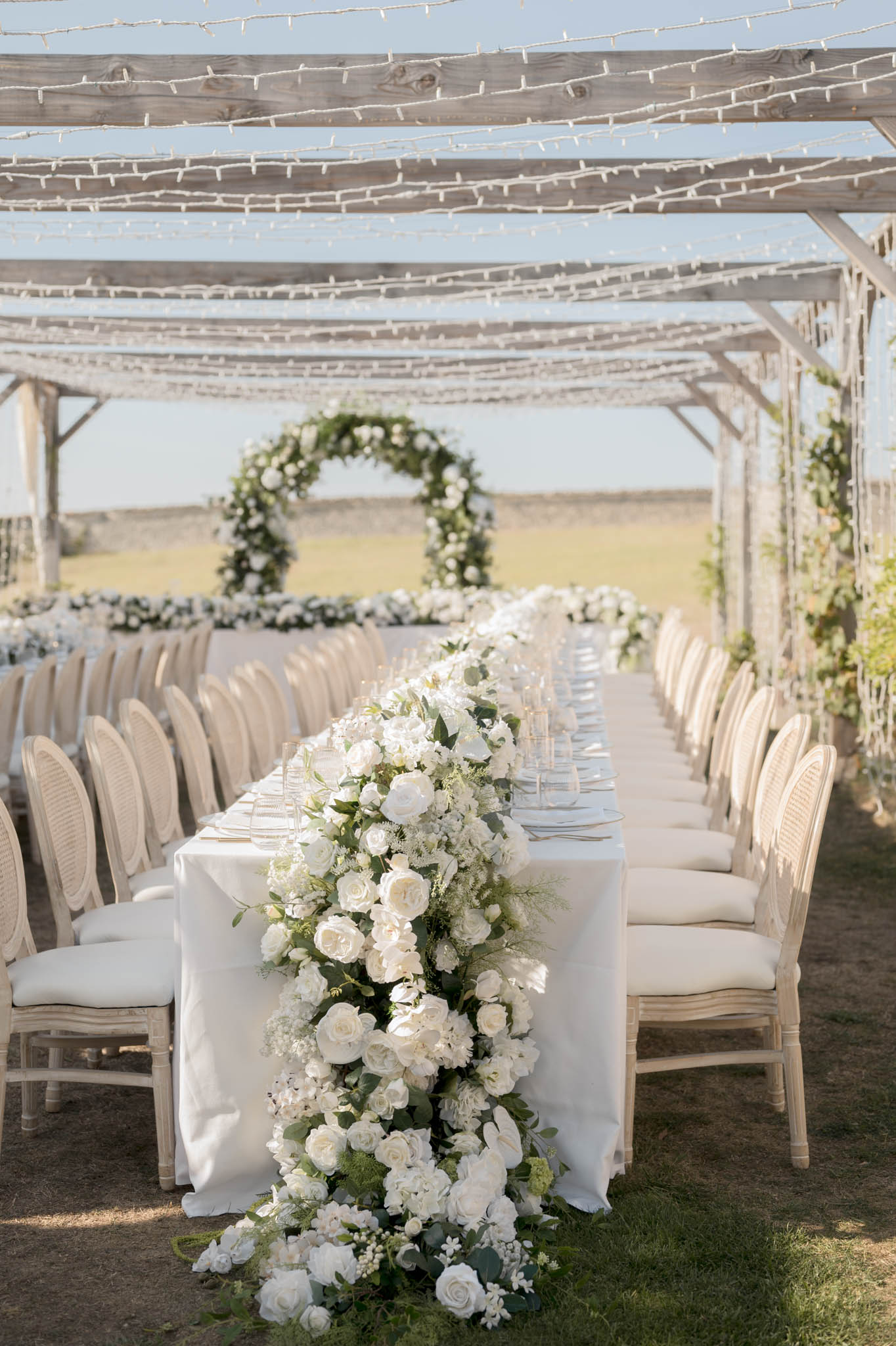 Long table with cascading white rose runner under fairy-lit pergola beside circular greenery arch