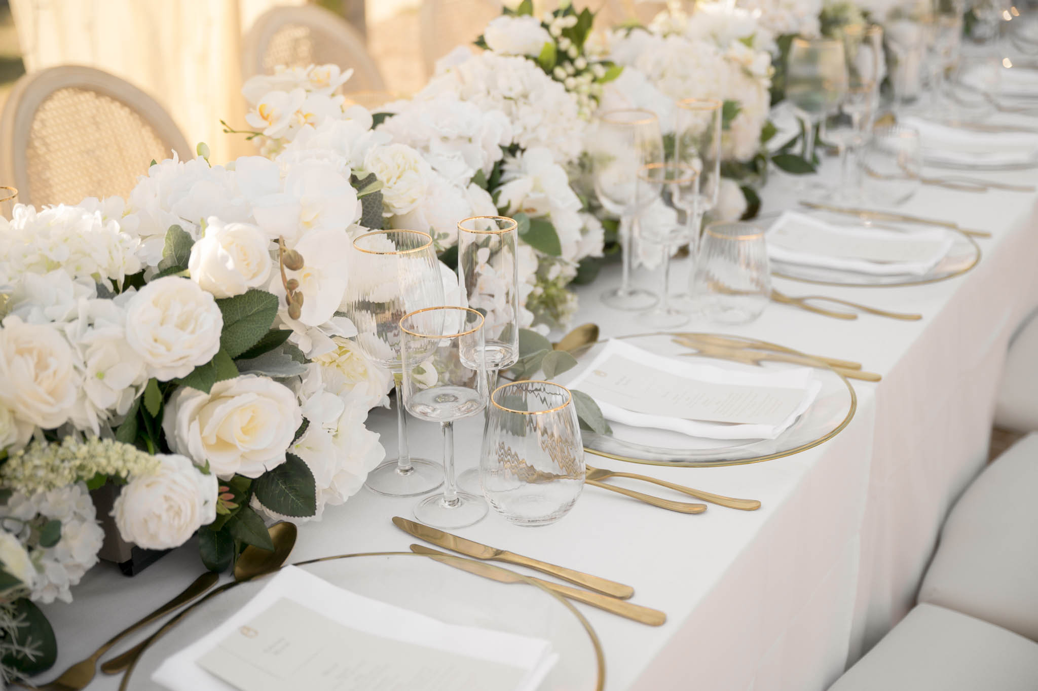 A close-up detail shot of a wedding reception tablescape, shot at a slight angle along the length of a long banquet table. The table is dressed with a white linen and features a lush low floral runner composed of white garden roses, white hydrangeas, white orchids, and eucalyptus foliage. Each place setting includes a clear glass charger plate with a thin gold rim, white folded napkins with printed menus, matte gold flatware, and a set of three ribbed glassware pieces — a wine glass, champagne flute, and stemless-style glass — each with a gold-rimmed edge. The chairs visible on both sides are upholstered in cream or ivory fabric, with at least one rattan-back chair visible on the left. The overall palette is white, ivory, and gold with green accents, consistent with a classic, all-white formal reception style. No people are present in the image.