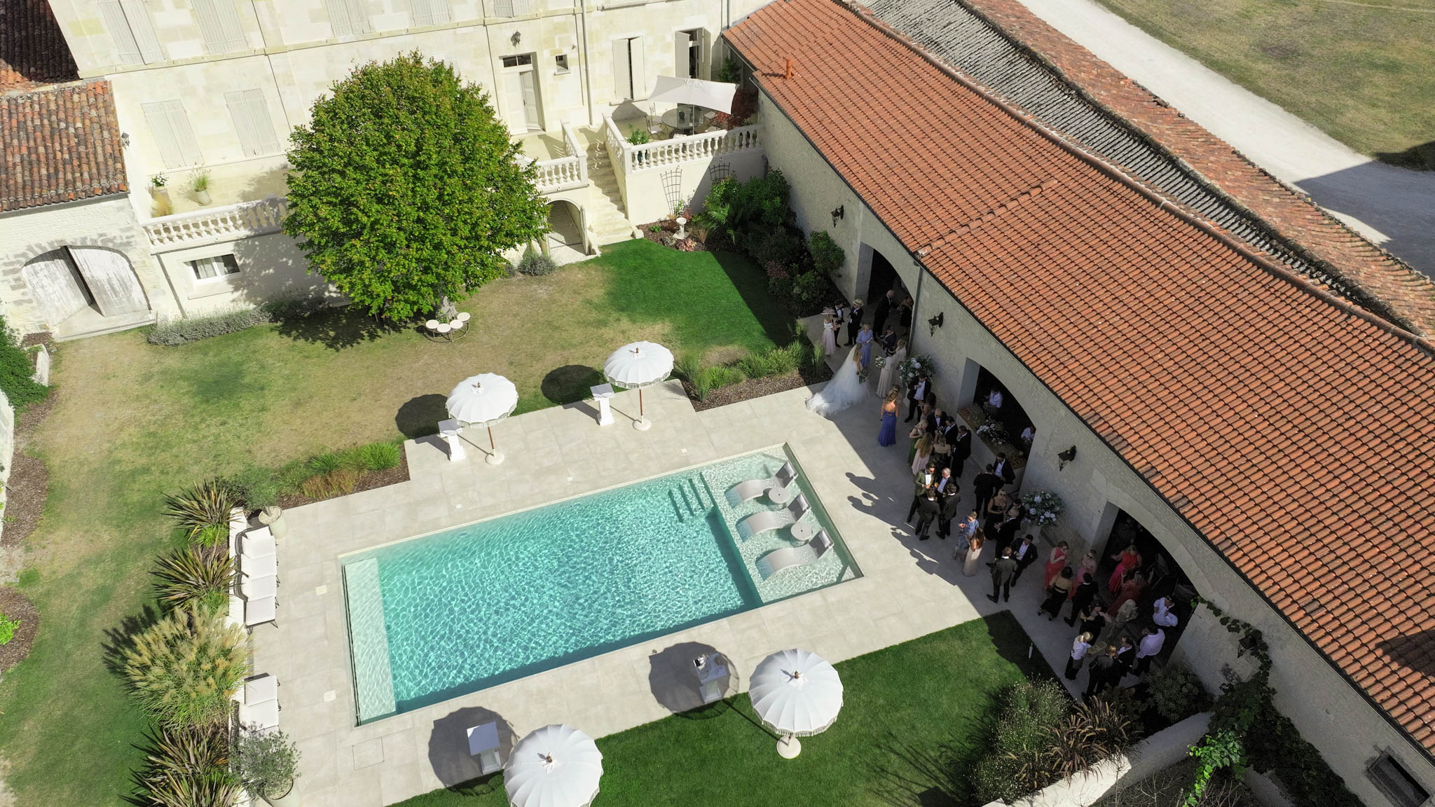 An aerial drone shot captures a cocktail hour in progress at a French château or domaine, with approximately 30–40 guests gathered along an outdoor covered arcade with terracotta roof tiles. A bride in a white gown is visible among the group near the entrance to the covered space, which is decorated with white floral arrangements. The property features a rectangular swimming pool with integrated stepped water features, surrounded by a light stone terrace dressed with white market umbrellas and sun loungers. The main château building, constructed in pale limestone with shuttered windows and a white balustrade balcony, forms the backdrop of a manicured inner courtyard. Potential venue feature image.