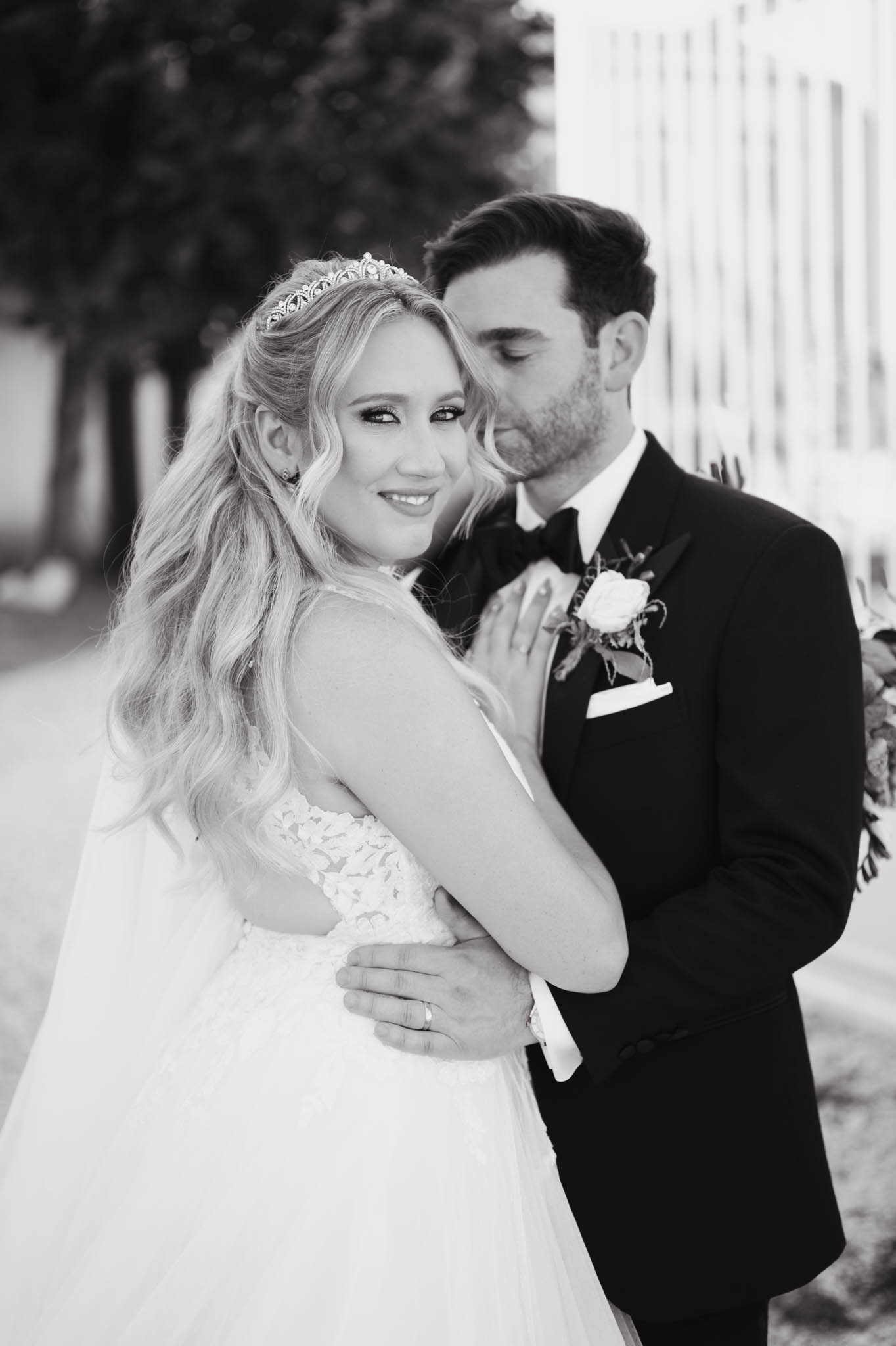 This is a black-and-white couple portrait taken outdoors, showing the bride and groom in a close embrace. The bride, facing the camera with long wavy blonde hair worn down, wears a lace-bodice ball gown with a tulle skirt and a delicate crystal tiara; the groom leans in toward her cheek with his eyes closed, wearing a dark tuxedo with a black bow tie, white pocket square, and a small rose boutonnière with greenery. The image is shot with strong contrast and soft background blur, giving the foreground subjects sharp definition against the light-toned architectural element and trees behind them. The composition is a mid-length portrait framing both figures from approximately the waist up.