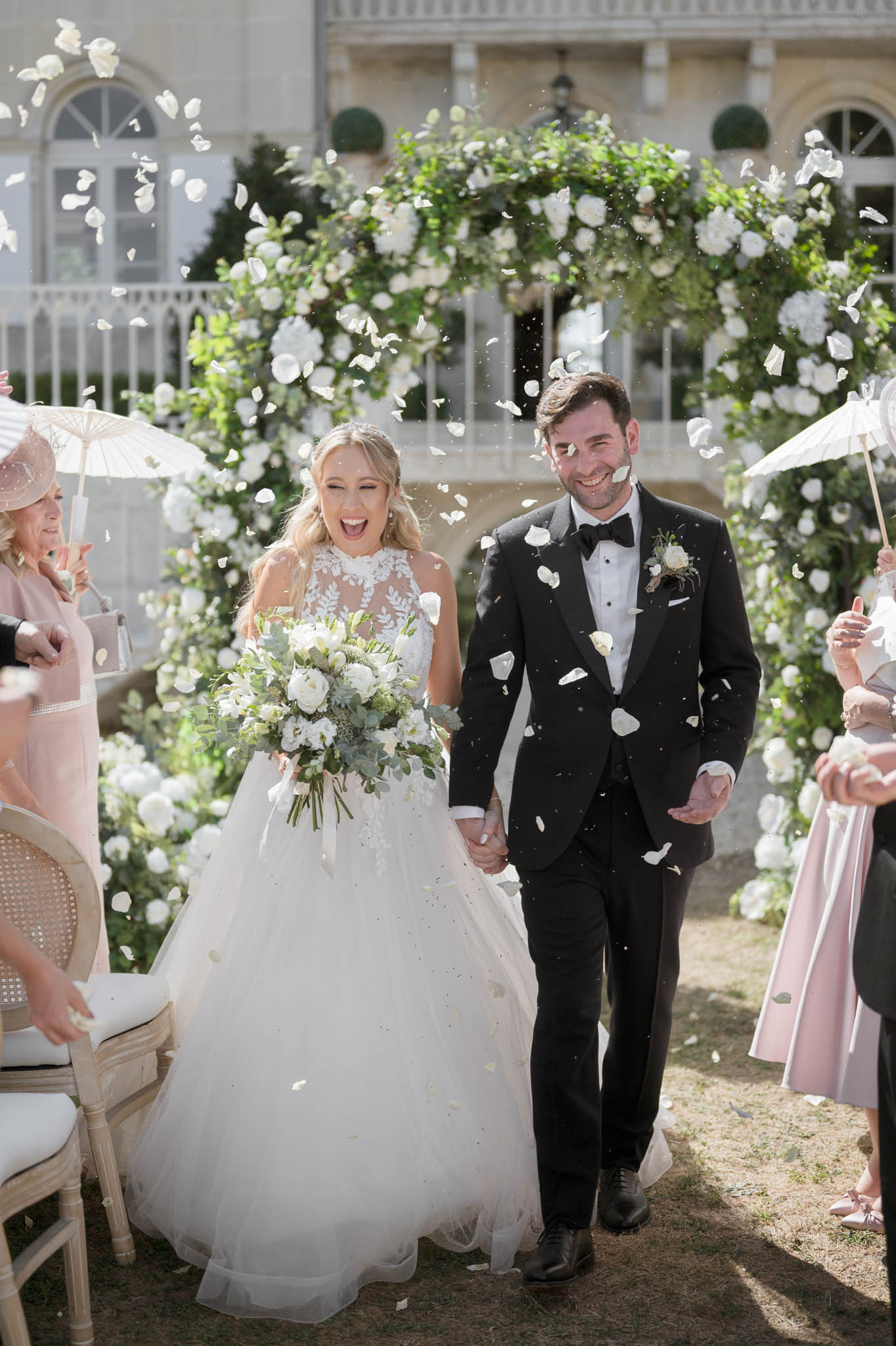 Bride and groom walk down aisle through white rose petal toss under circular floral arch at chateau ceremony