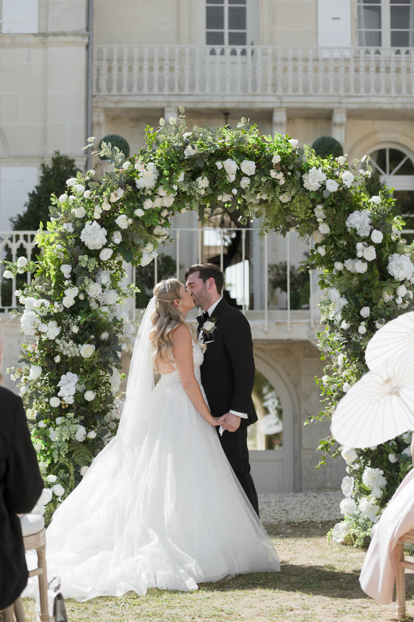 First kiss under circular white rose and hydrangea arch before chateau with wrought-iron balconies