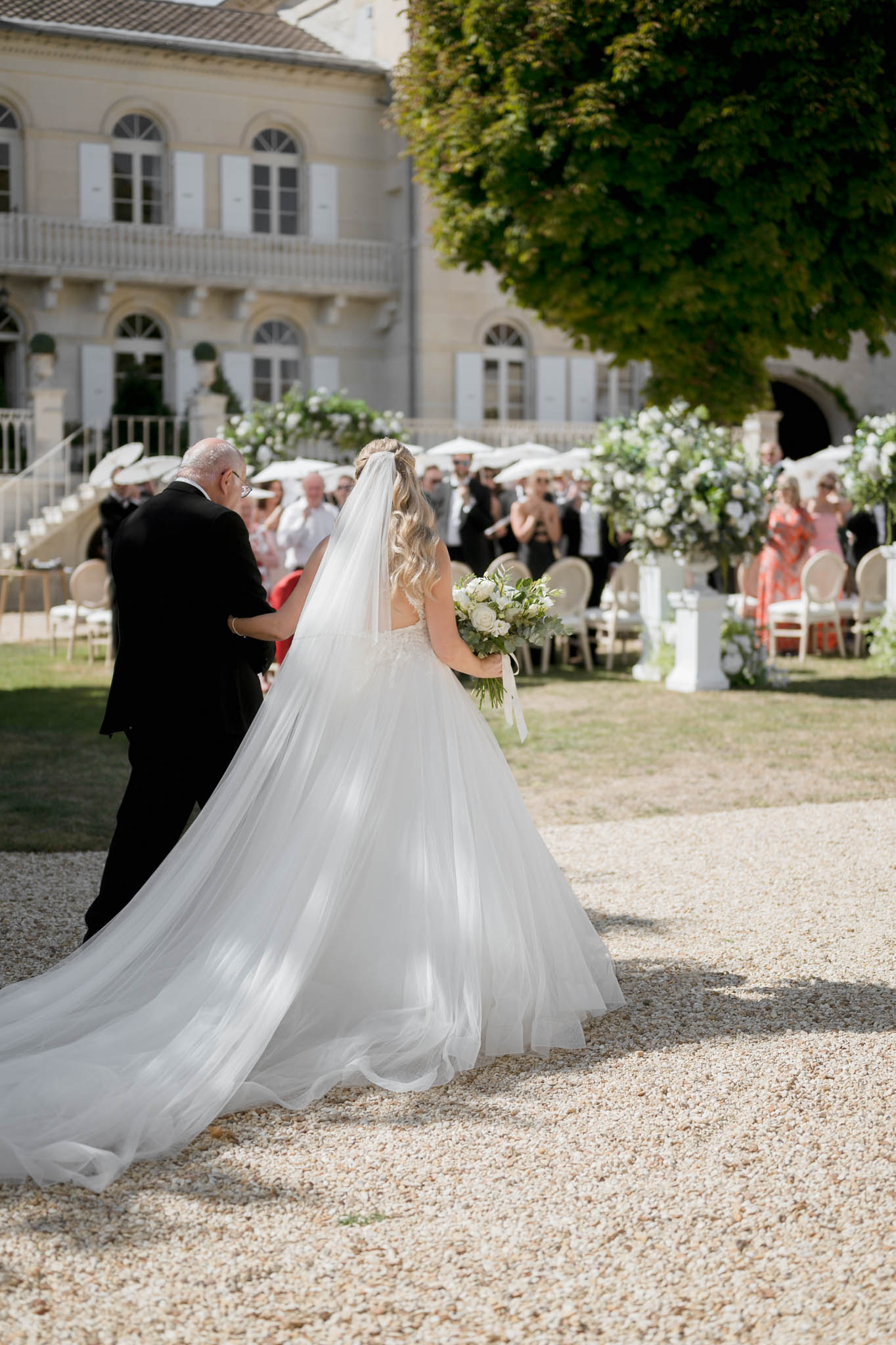 Bride with cathedral veil escorted down aisle past gold chairs white parasols and pedestal floral arrangements
