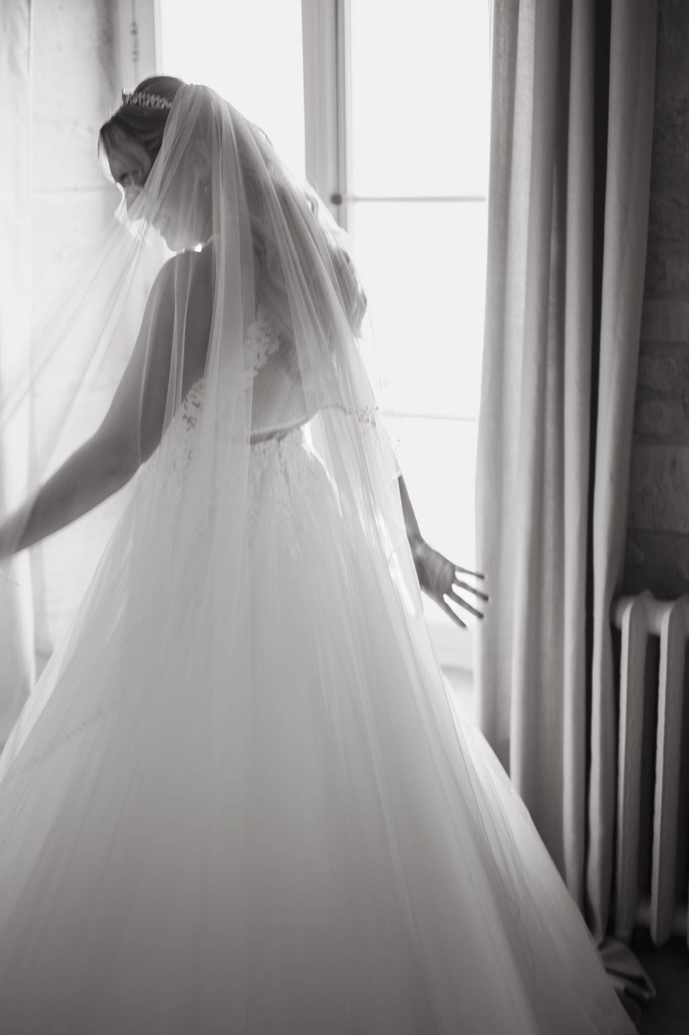 Bride in lace ballgown with tiara and two-tier cathedral veil settling into dress by window in B&W