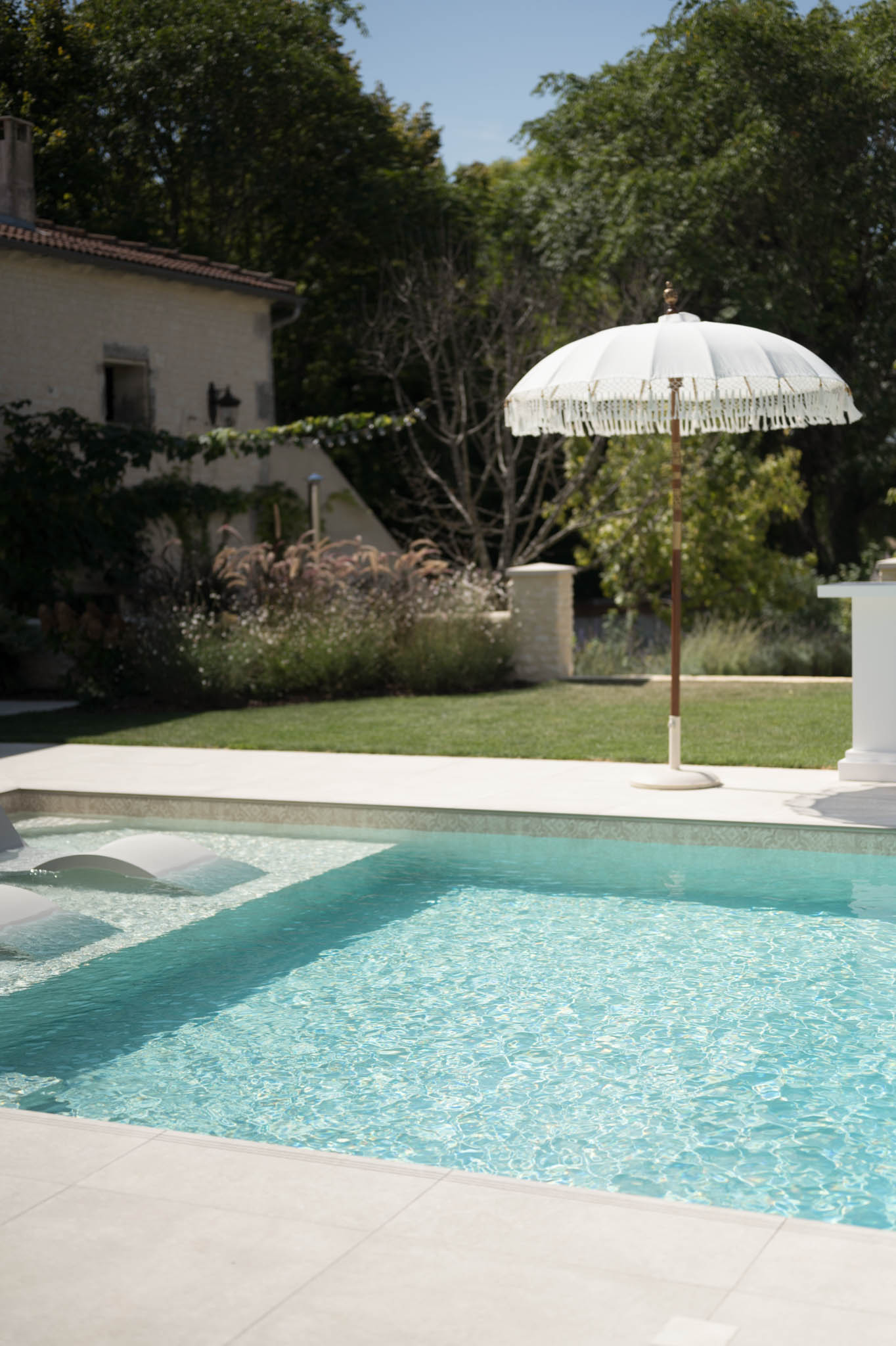 Turquoise pool with white fringed parasol and submerged lounge chairs at a French stone manor house