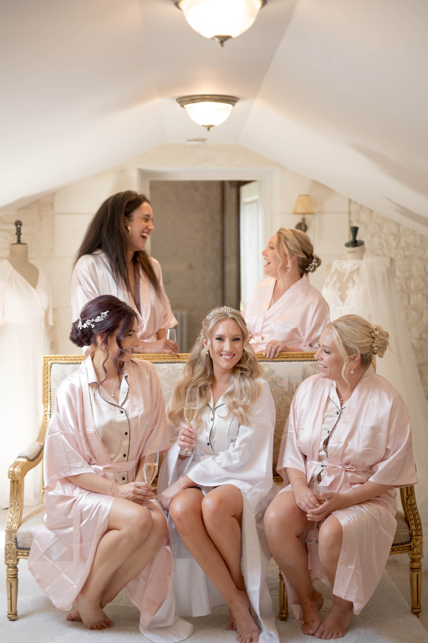 A getting-ready scene set in an indoor bridal suite with a vaulted white ceiling, lit by ceiling-mounted flush lights. Five women are gathered together — the bride sits centrally on a gold-framed settee wearing a white satin short robe with black piping trim and a crystal tiara, holding a champagne flute; her hair is styled in loose blonde waves. Four bridesmaids wear matching blush pink satin robes, two seated flanking the bride and two standing behind, all with upswept or styled hair and small hair accessories. All five women are holding or have glasses of champagne and are laughing and interacting naturally. In the background, white lace wedding dresses are displayed on dress forms and hang on the walls, and a large gold-framed mirror reflects part of the room. The composition is a wide group portrait shot from a slightly low angle.