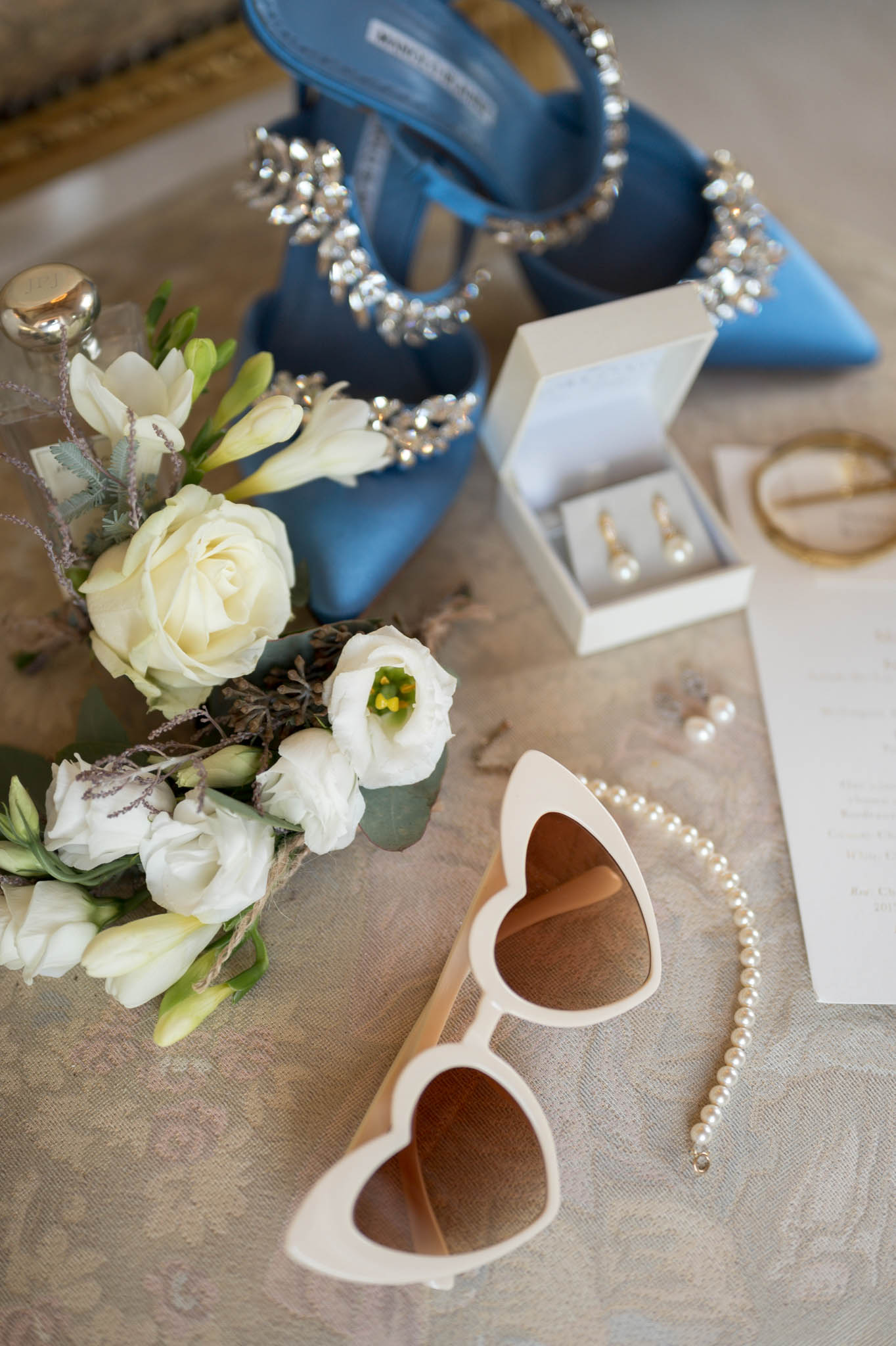 Bridal flat lay with blue satin heels, ivory rose bouquet, pearl jewelry, gold earrings and sunglasses
