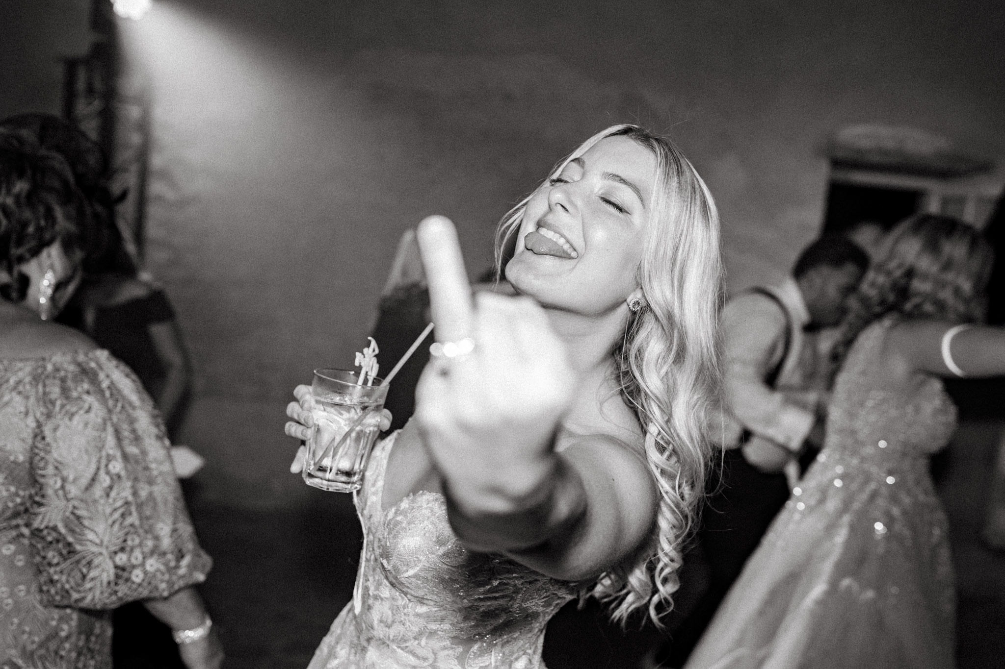 Black-and-white close-up portrait of a female wedding guest dancing at an indoor reception. She has long wavy blonde hair, is wearing a lace or embroidered dress, and is holding a cocktail glass with a straw in one hand while extending her other hand toward the camera with her middle finger raised. Her eyes are partially closed and her tongue is out in a playful expression. The background shows several other guests dancing under a spotlight in what appears to be a stone-walled venue space, with atmospheric haze visible in the upper portion of the frame. The image has high contrast with bright highlights on the subject and darker mid-tones in the background. The shot is a candid, close-up portrait taken at dance floor level.