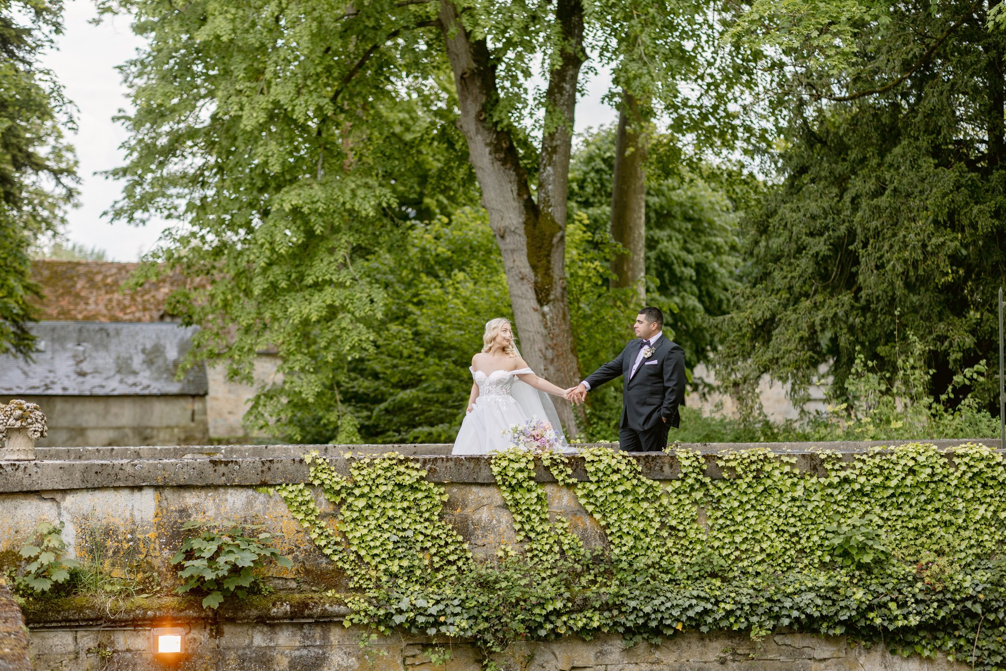 A bride and groom stand on a wide stone terrace or bridge covered in ivy, holding hands during an outdoor couple portrait. The bride wears an off-the-shoulder white ballgown with lace appliqué bodice and carries a loose bouquet with pink, lavender, and yellow blooms. The groom is dressed in a dark navy suit with a light pink tie and a floral boutonnière. A stone outbuilding with a slate roof is visible to the left, and mature trees frame the background. The shot is taken from a low, wide angle, with the ivy-covered stone wall filling the lower half of the frame.