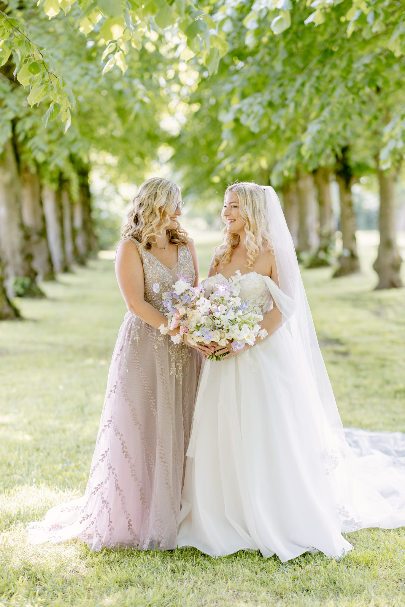An outdoor portrait of a bride and one companion — likely a bridesmaid or mother of the bride — standing together in a tree-lined avenue, facing each other and smiling. The bride wears a strapless ivory ballgown with a long cathedral veil and loose blonde curls, and holds a loose, garden-style bouquet featuring white sweet peas, soft blush roses, lavender blooms, and cream florals. Her companion wears a floor-length blush-taupe gown with embroidered leaf detailing and a V-neckline, and also holds a smaller version of the same floral arrangement. The overall styling is romantic and soft, with a pastel floral palette of white, blush, and lilac. The shot is a full-length portrait taken in natural daylight with a bright, airy feel.