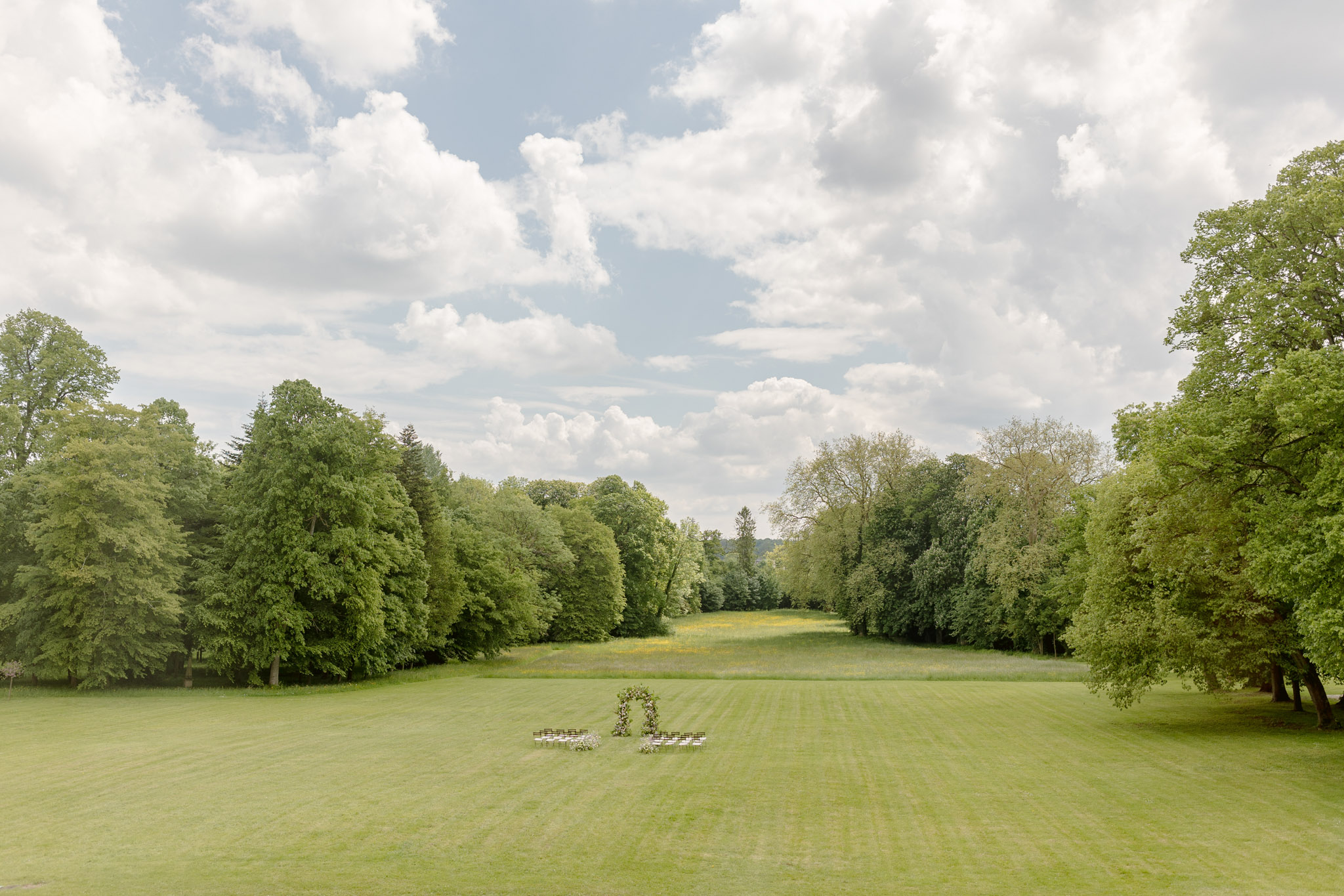 Aerial view of outdoor ceremony setup with floral arch and white chairs on manicured lawn between tall trees