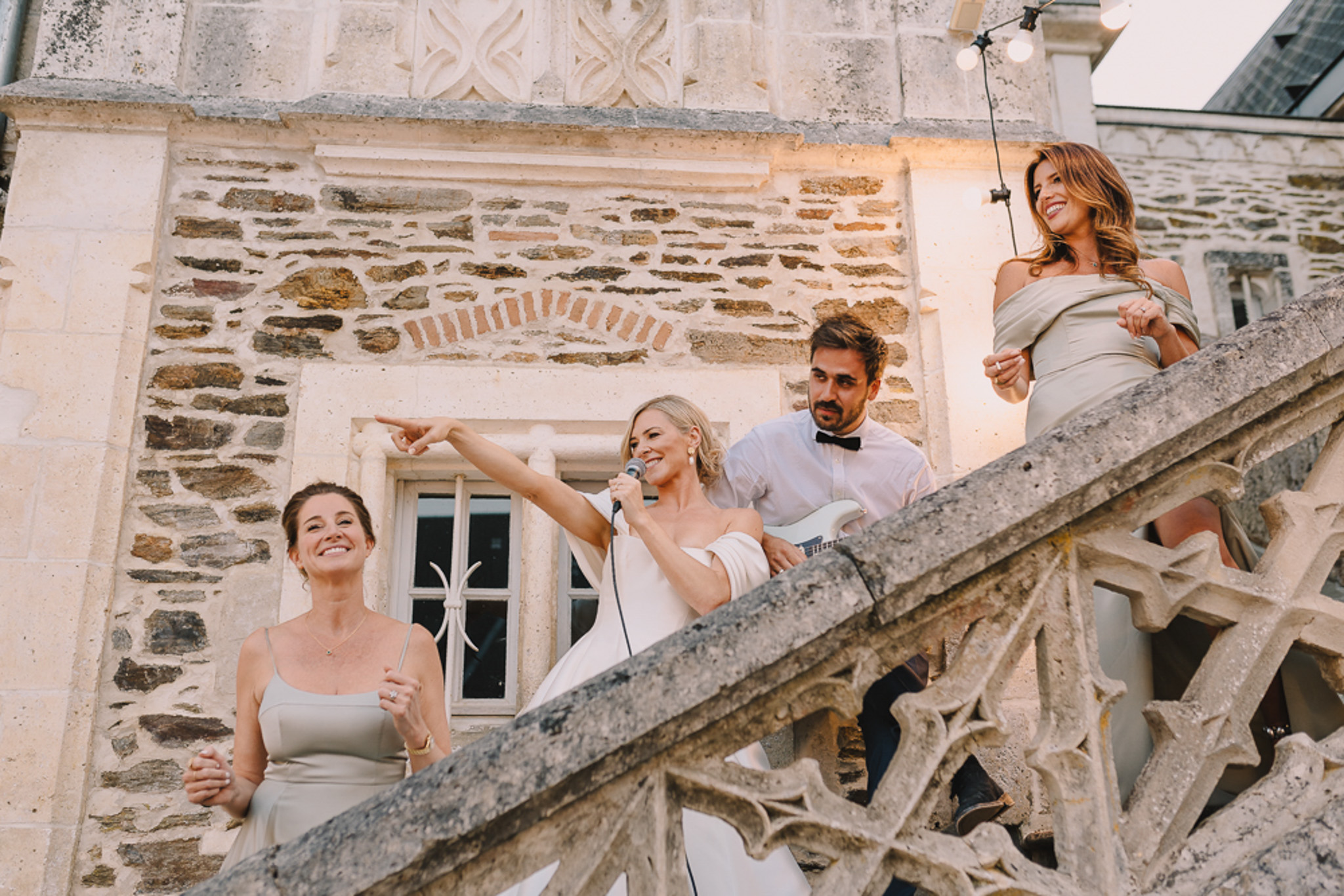 Bride singing into microphone on chateau steps with guitarist and sage-dressed bridesmaids under festoon lights