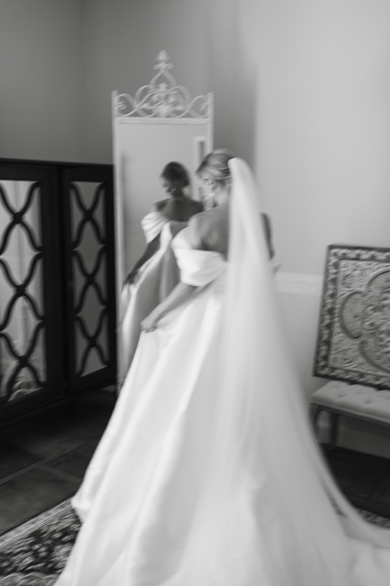 A black-and-white getting-ready image of a bride standing in front of an ornate white-framed floor mirror in an indoor room, with her reflection visible. The bride wears a full-skirted ball gown with off-the-shoulder or detachable puff sleeves and a long veil, with her hair styled in an updo. The image is intentionally motion-blurred, giving the scene a sense of movement as the bride adjusts her dress or sleeves. The room features a decorative lattice-patterned door or screen on the left, a patterned area rug, and an ornate carved bench or chair visible to the right. The composition is a mid-range portrait with strong contrast between the bright white of the gown and veil and the darker room elements.