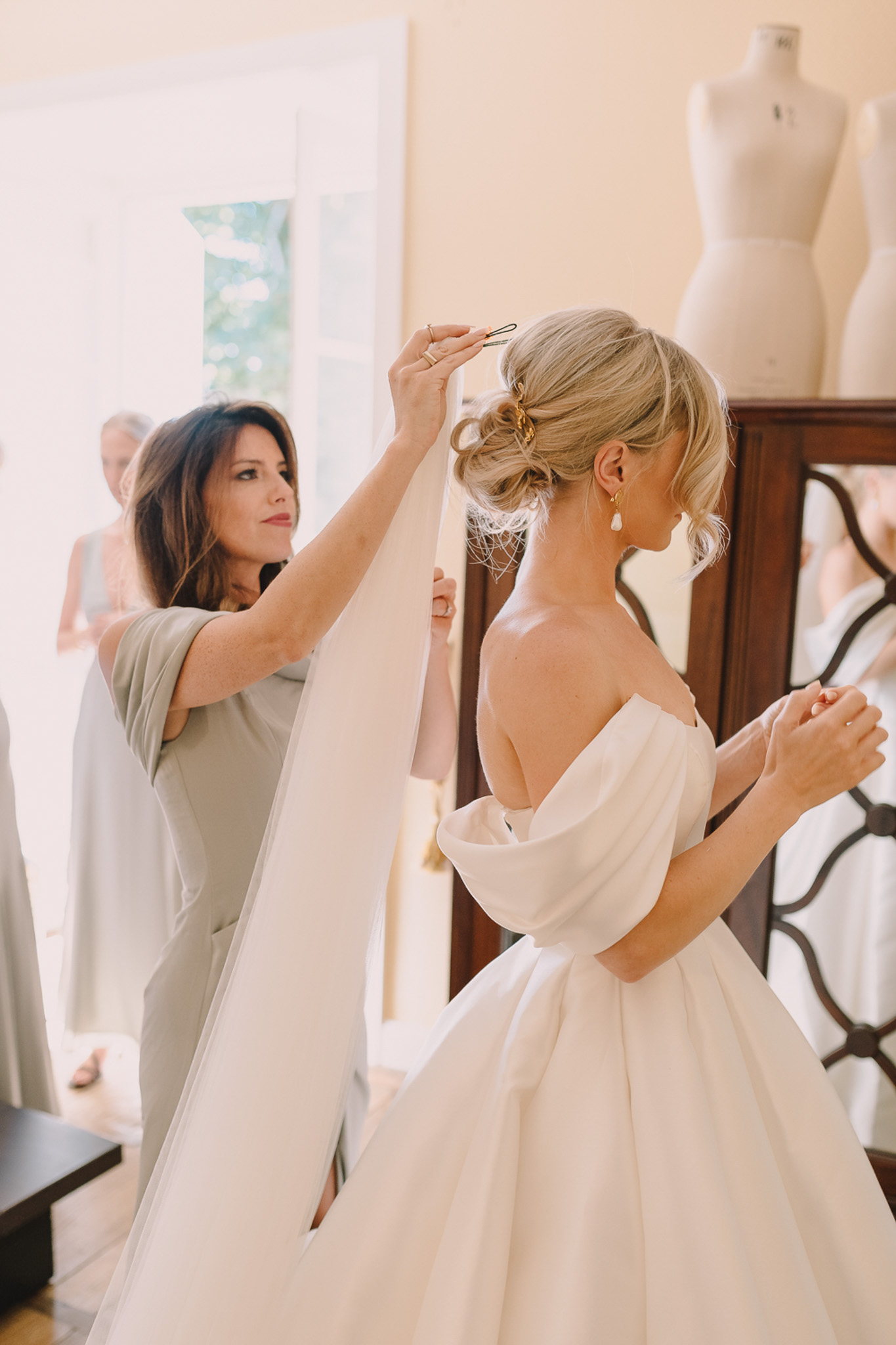 Bridesmaid in sage green dress pinning ivory veil into bride's updo showing open-back ball gown and gold earrings