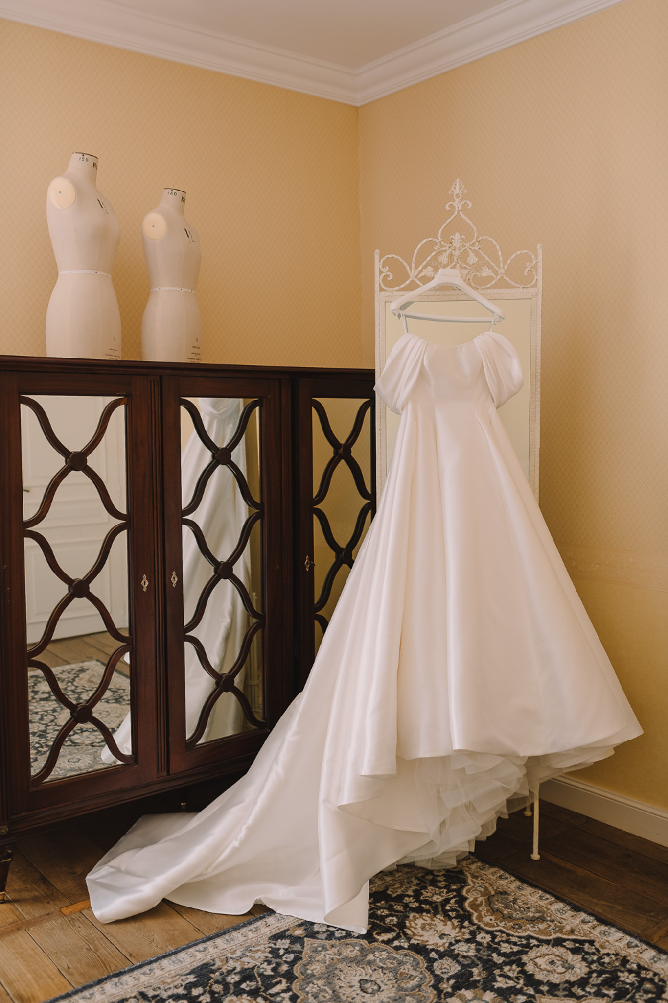 A getting-ready detail shot of an ivory satin wedding gown hung on a white ornate wrought-iron dress stand in an interior room. The gown features off-the-shoulder puff sleeves and a full ball gown skirt with a long train that pools on the hardwood floor. The room has warm yellow walls, white crown molding, and a dark mahogany mirrored wardrobe with lattice-pattern doors, in which the dress is partially reflected. Two white dressmaker mannequin torsos sit atop the wardrobe. A navy and cream patterned area rug covers part of the floor. The composition is a full-length vertical portrait-style shot capturing the entire dress and its surroundings.