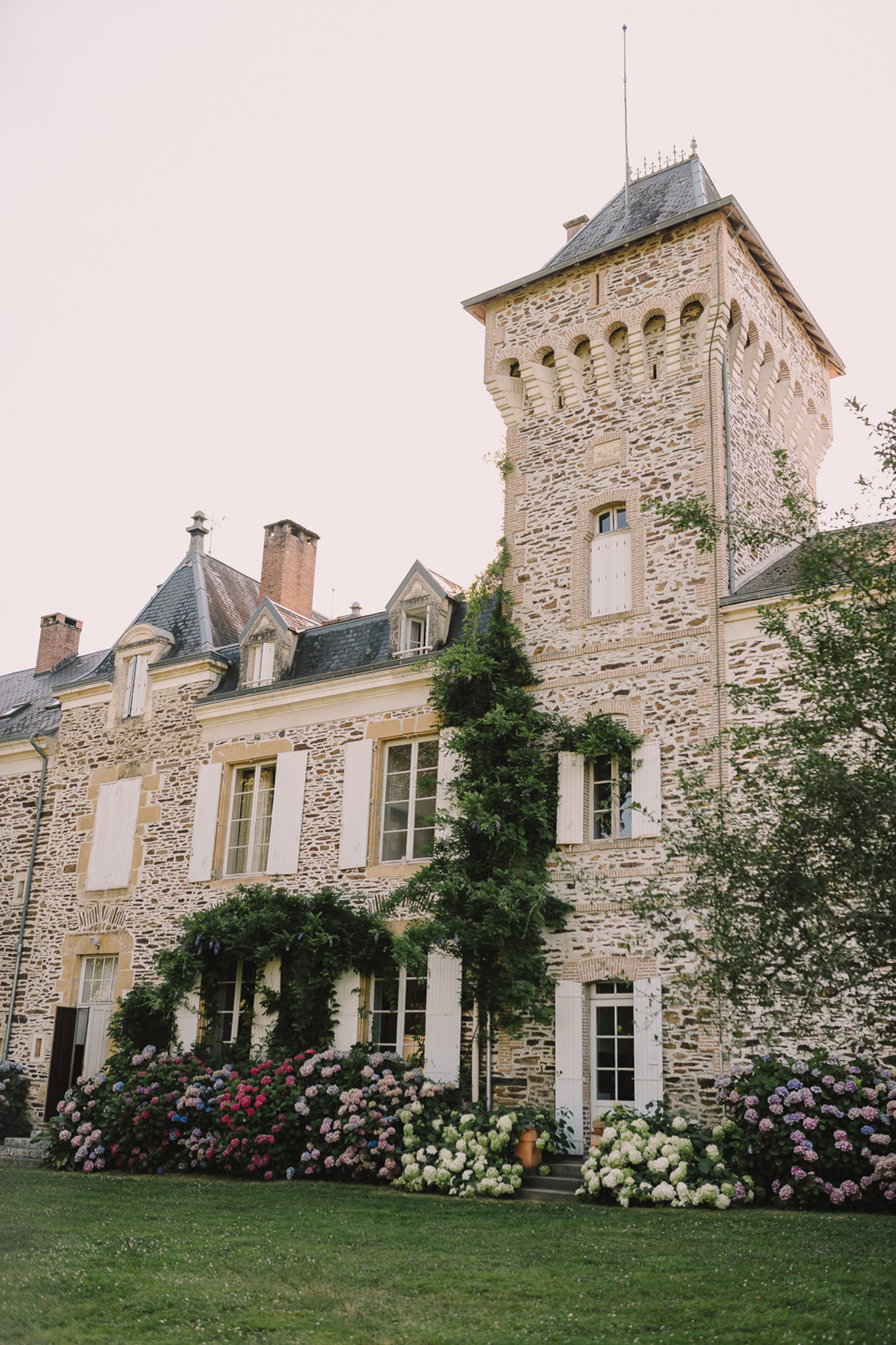 French chateau exterior with square stone tower, slate roof, white shutters, and hydrangea bushes in bloom