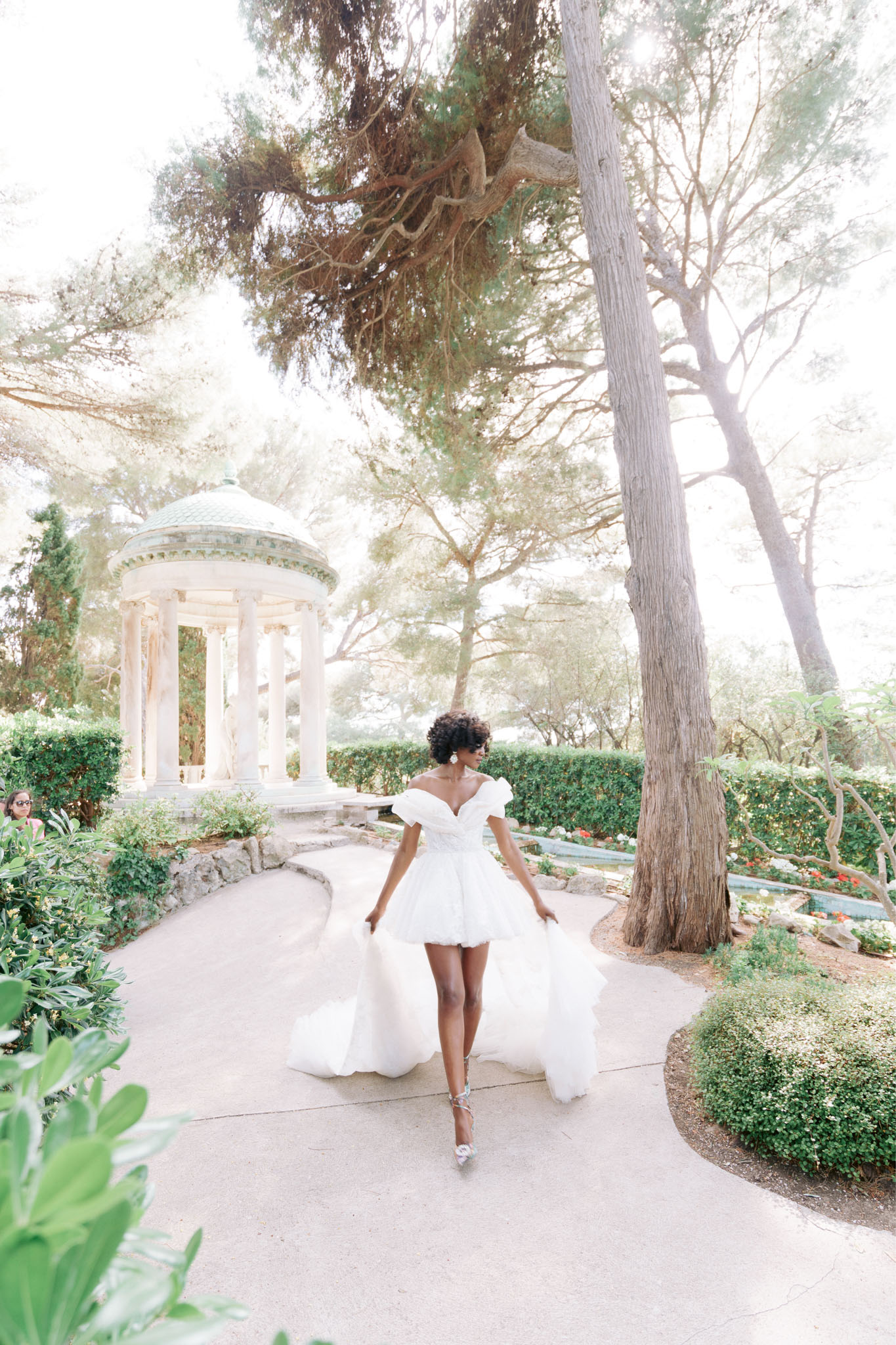 Bride walking away twirling white off-shoulder tulle mini dress with train past neoclassical rotunda in formal garden
