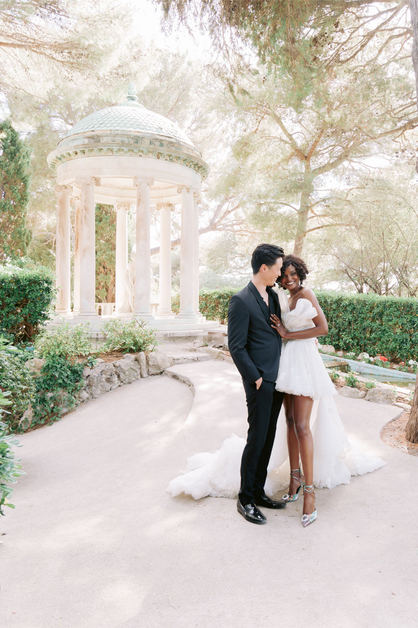 Couple in mini dress with tulle train and black suit smile on gravel path before neoclassical rotunda