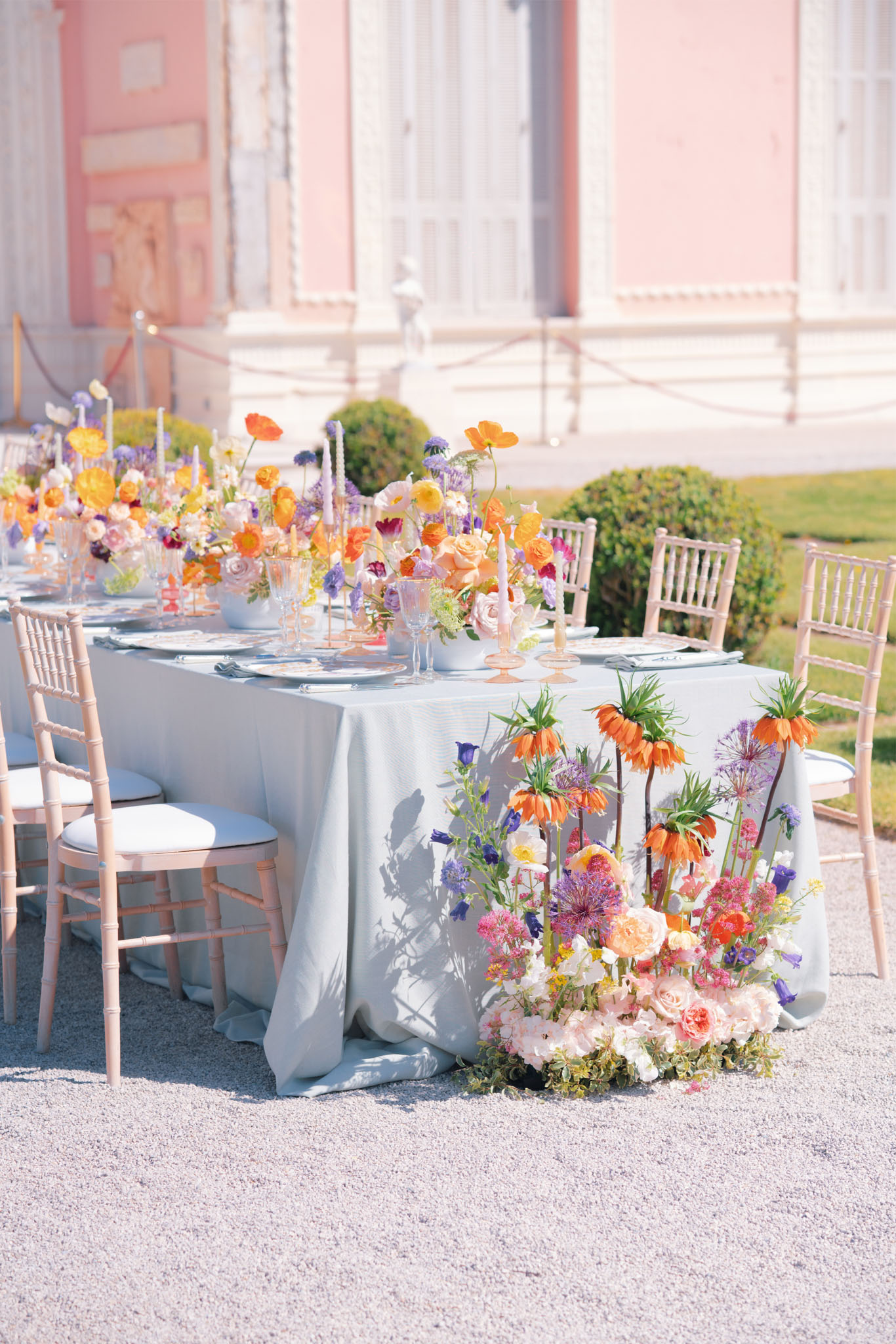 An outdoor reception table setup photographed in wide shot on a gravel courtyard in front of a pink neoclassical building with white architectural detailing. The long rectangular table is dressed in a pale blue-grey linen tablecloth and surrounded by blush pink chiavari chairs with white cushions. The tablescape features an abundant floral runner composed of orange Iceland poppies, peach and blush garden roses, lavender scabiosa, purple alliums, yellow ranunculus, and mixed wildflowers, with slender lilac and peach taper candles interspersed throughout. At the base of the table end sits a large floor-level floral installation including orange fritillaria imperialis (crown imperial), purple alliums, blush roses, hot pink blooms, and trailing greenery. Place settings include white plates with chargers, sage green cloth napkins, and amber-tinted glassware. The overall color palette combines orange, peach, lavender, blush, and yellow in a maximalist garden-party style. Potential venue feature image.