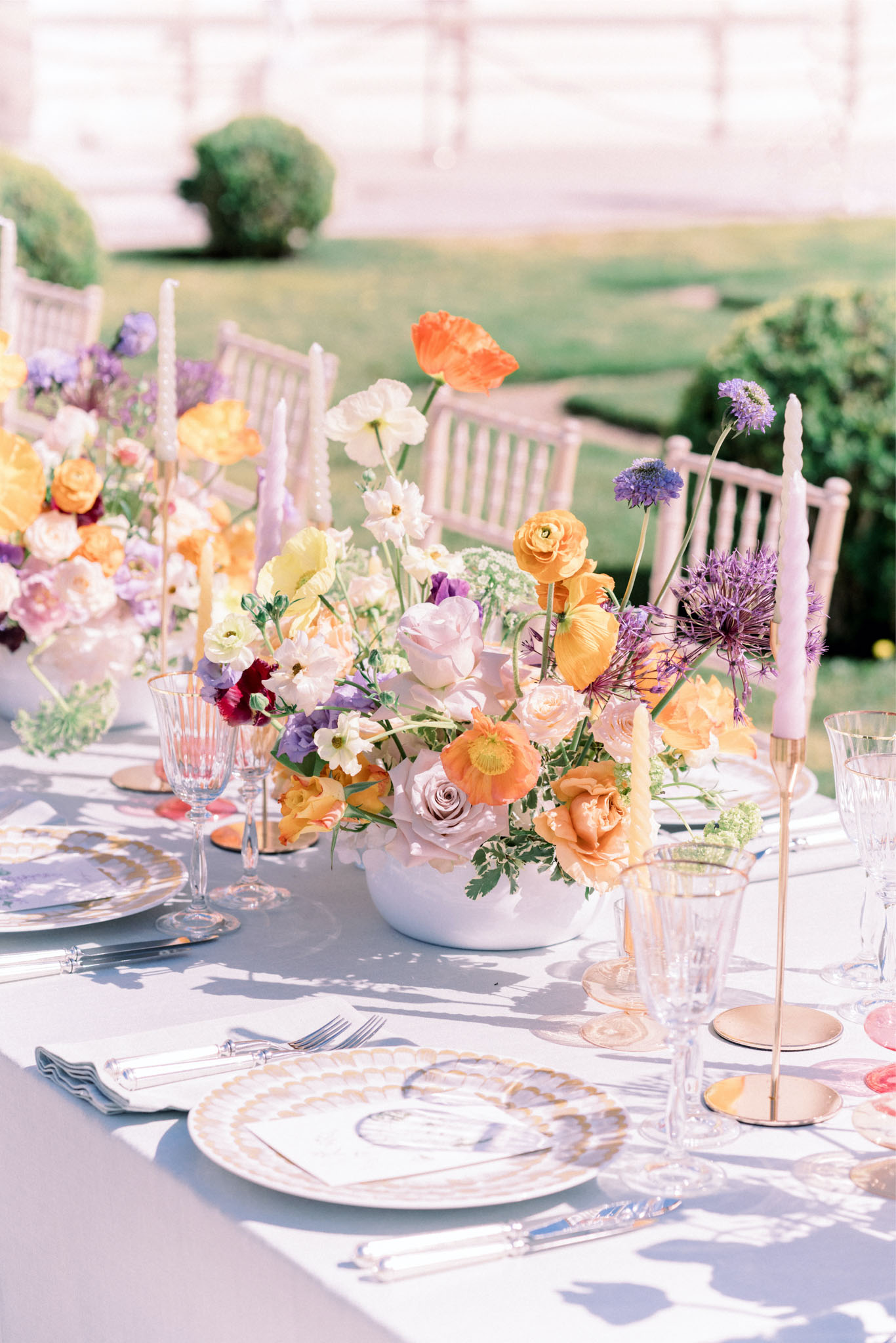 A close-up detail shot of an outdoor wedding reception tablescape set in a formal garden setting with manicured hedges visible in the background. The table is dressed with a pale grey linen and features elaborate floral centerpieces arranged in white ceramic bowls, filled with orange and peach poppies, blush and lavender roses, yellow ranunculus, white cosmos, deep burgundy blooms, purple allium, and delicate white filler flowers. Tall tapered candles in lavender and pale yellow shades are placed in gold candlestick holders throughout the table. Place settings include gold-rimmed decorative charger plates with an illustrated motif, crystal glassware in clear and blush pink tones, and silver flatware with acrylic handles. The overall decor palette combines warm peach and orange tones with soft lilac and lavender accents against the grey linen, creating a colorful, garden-forward aesthetic. White chiavari chairs are visible in the background.
