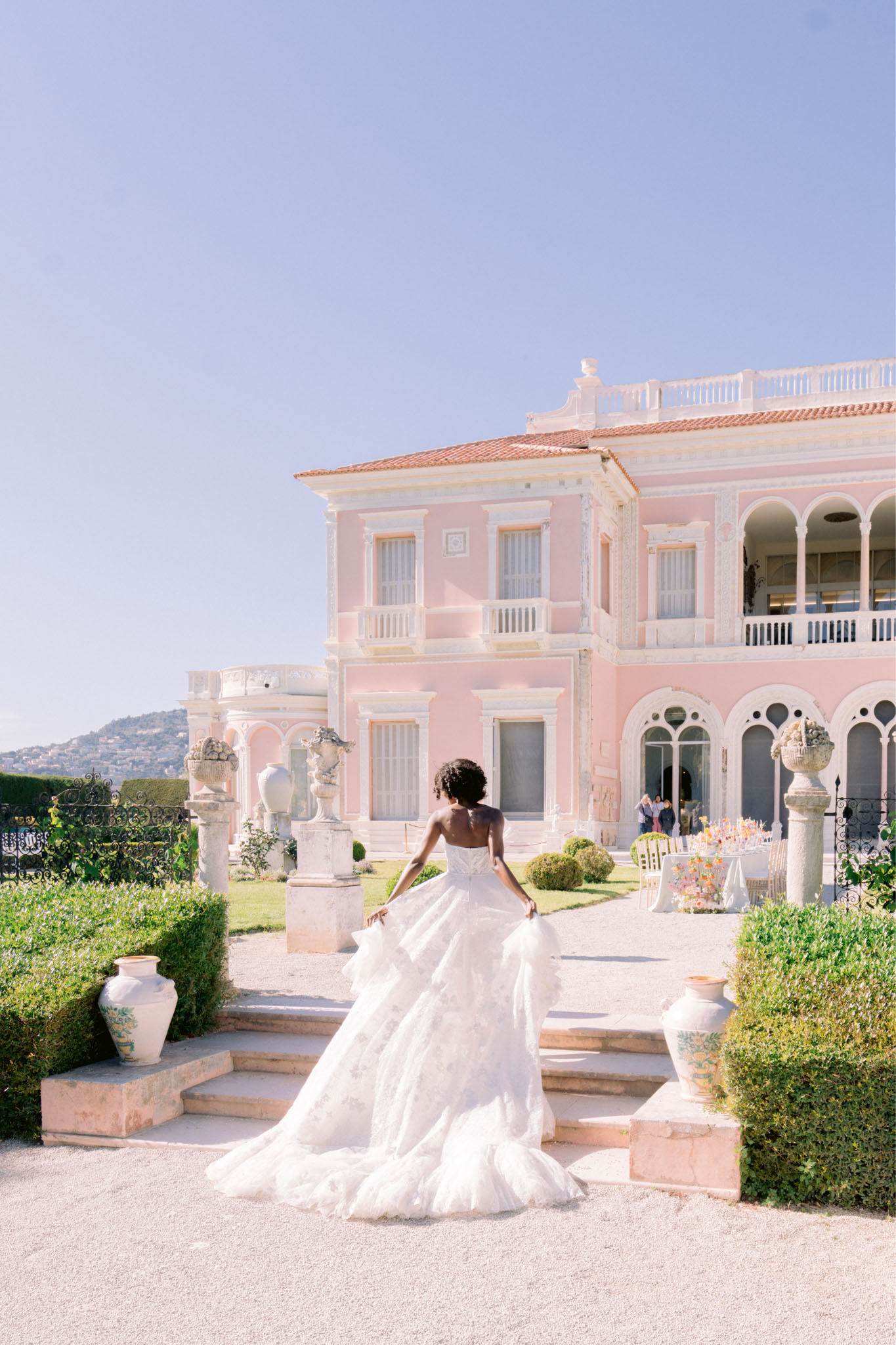 Bride from behind ascending steps of pink Belle Epoque villa in tiered tulle ballgown with open back