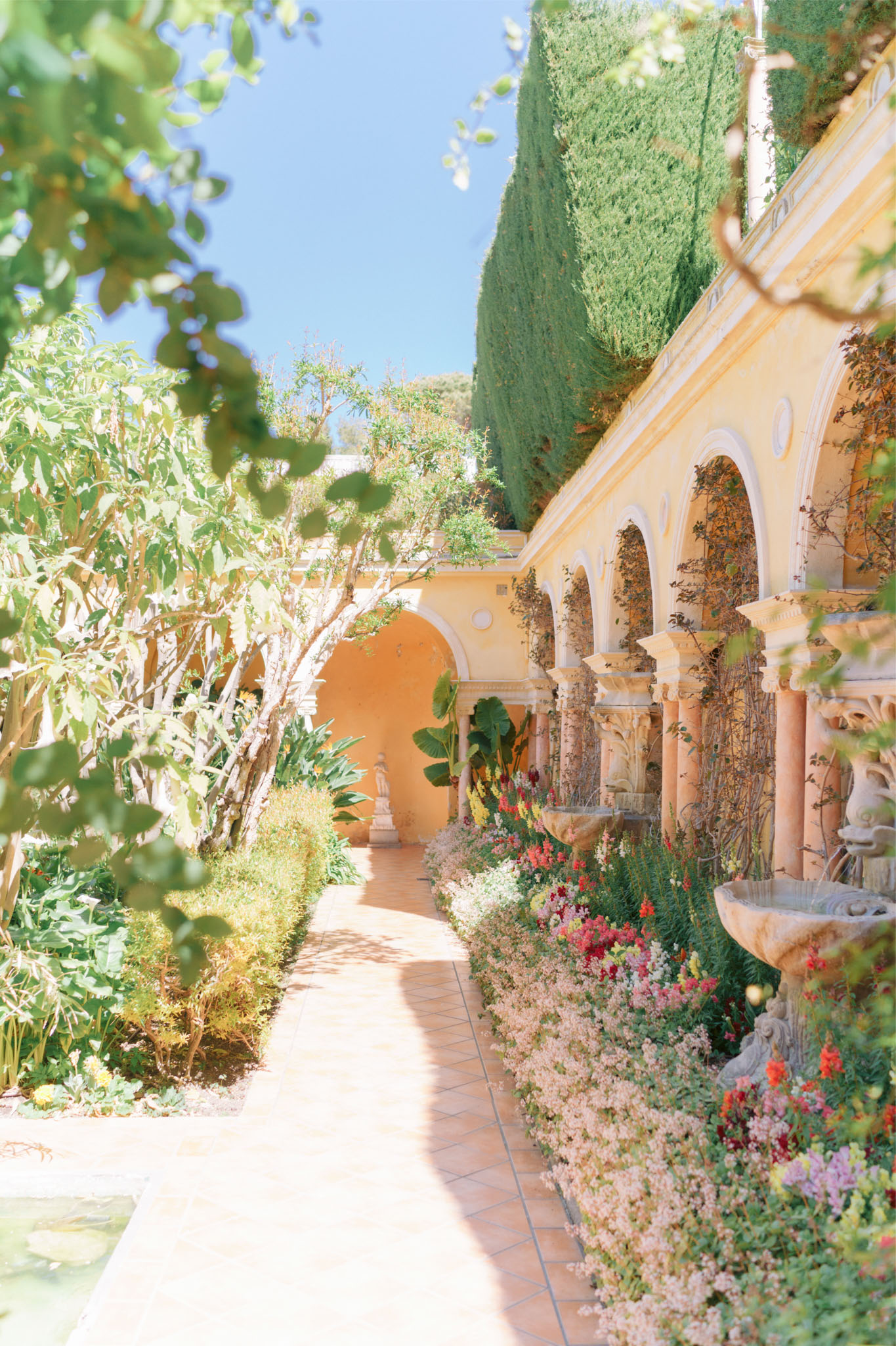 A wide shot of an outdoor venue corridor featuring a warm yellow ochre building with a series of ornate white-trimmed arches supported by stone columns, lined with a terracotta-tiled walkway. Dense floral plantings run along the base of the arched colonnade, including blush pink, coral, and deep red blooms — appearing to include snapdragons, alyssum, and similar garden flowers. A carved stone wall fountain is visible on the right side. No people are present in the image. The overall aesthetic is Mediterranean or Spanish Colonial in style, with a classic, garden-forward character. Potential venue feature image.