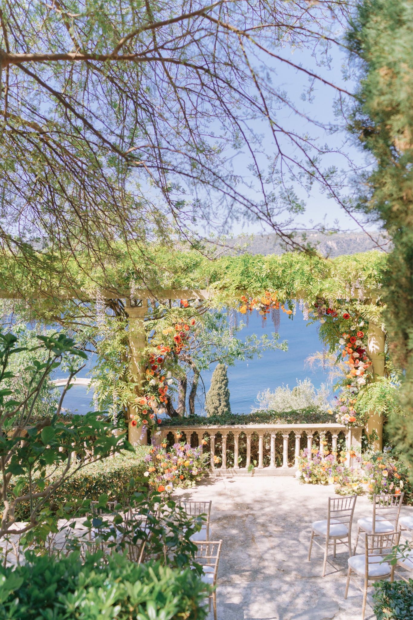 An outdoor wedding ceremony setup photographed from a distance through tree branches, framing a terrace with a stone balustrade overlooking a blue body of water and hillside beyond. The ceremony space features a large floral arch adorned with vibrant orange, red, and yellow blooms alongside trailing lavender wisteria and lush greenery, positioned at the far end of a stone-paved aisle. Low floral arrangements in matching orange, yellow, lavender, and blush tones line both sides of the aisle. Gold chiavari chairs with white cushions are arranged in rows on either side of the aisle, and no guests are present, indicating this is a pre-ceremony setup shot. The overall floral palette is bold and colorful with a garden-party aesthetic. Wide establishing shot taken from an elevated vantage point partially obscured by surrounding trees and foliage. Potential venue feature image.