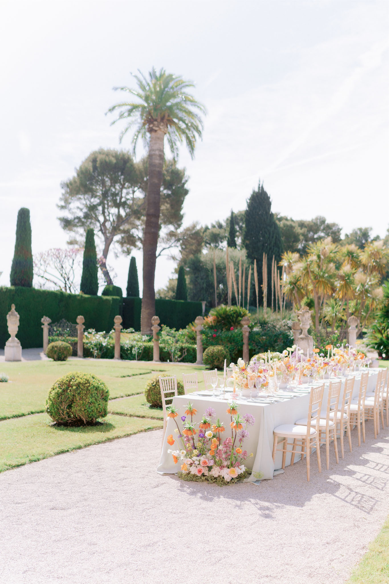 An outdoor wedding reception table is set up on a gravel path within a formal garden featuring clipped hedges, stone balustrade pillars, topiary spheres, and tall cypress and palm trees. The long feasting table is dressed in a cream linen and lined with natural wood chiavari chairs with ivory cushions. A dramatic low floral installation runs along the table and cascades down the end closest to the camera, featuring orange fritillaria, lavender alliums, peach and blush garden roses, and yellow blooms in a wildflower-style arrangement with a moss base. Slim taper candles in soft yellow and ivory tones are interspersed along the table runner, alongside glassware and sage-green napkins. The wide-angle shot captures the full length of the table against the structured garden setting, suggesting a South of France venue. Potential venue feature image.