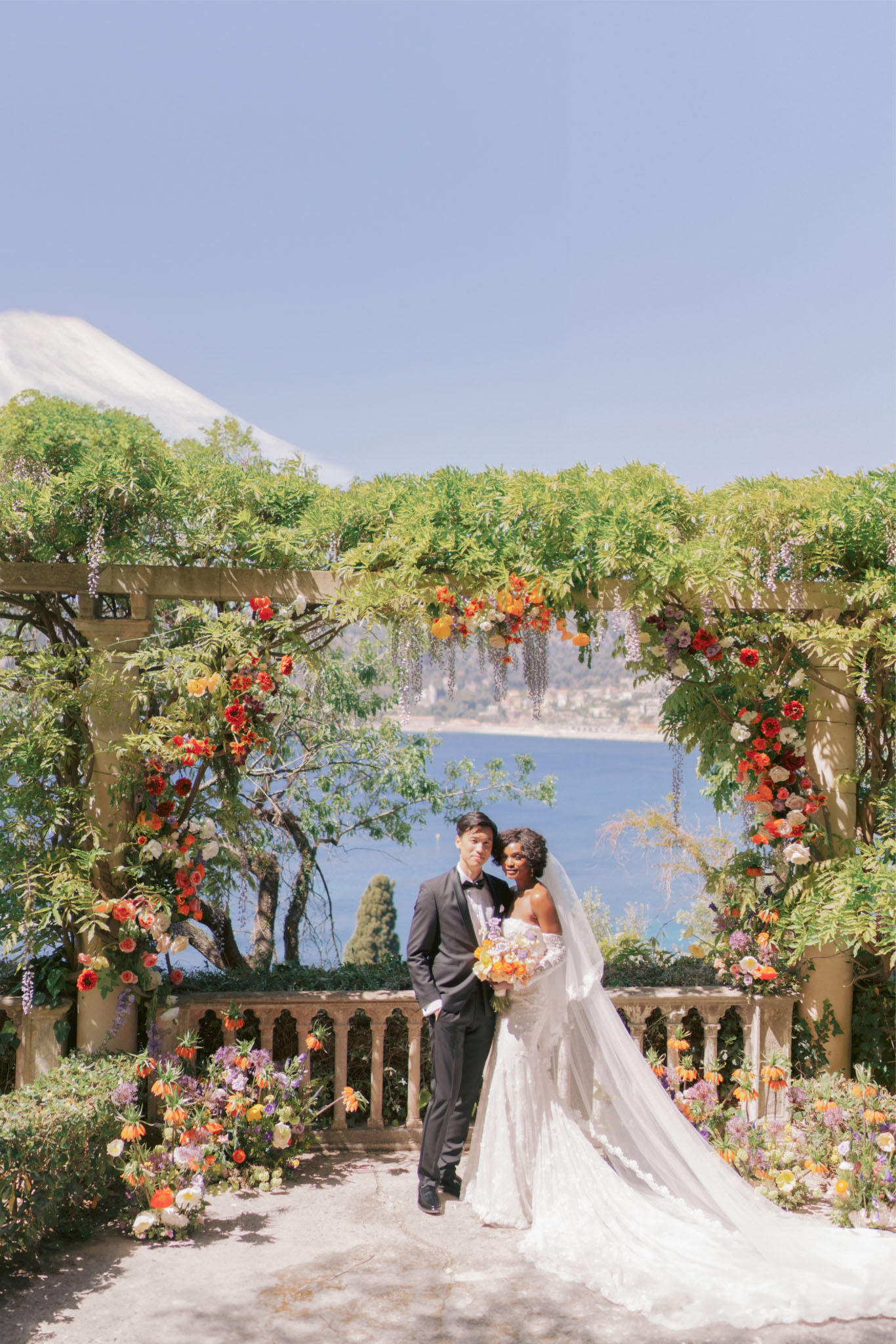 Couple at wisteria terrace with maximalist warm-toned floral arch overlooking Mediterranean sea and coast