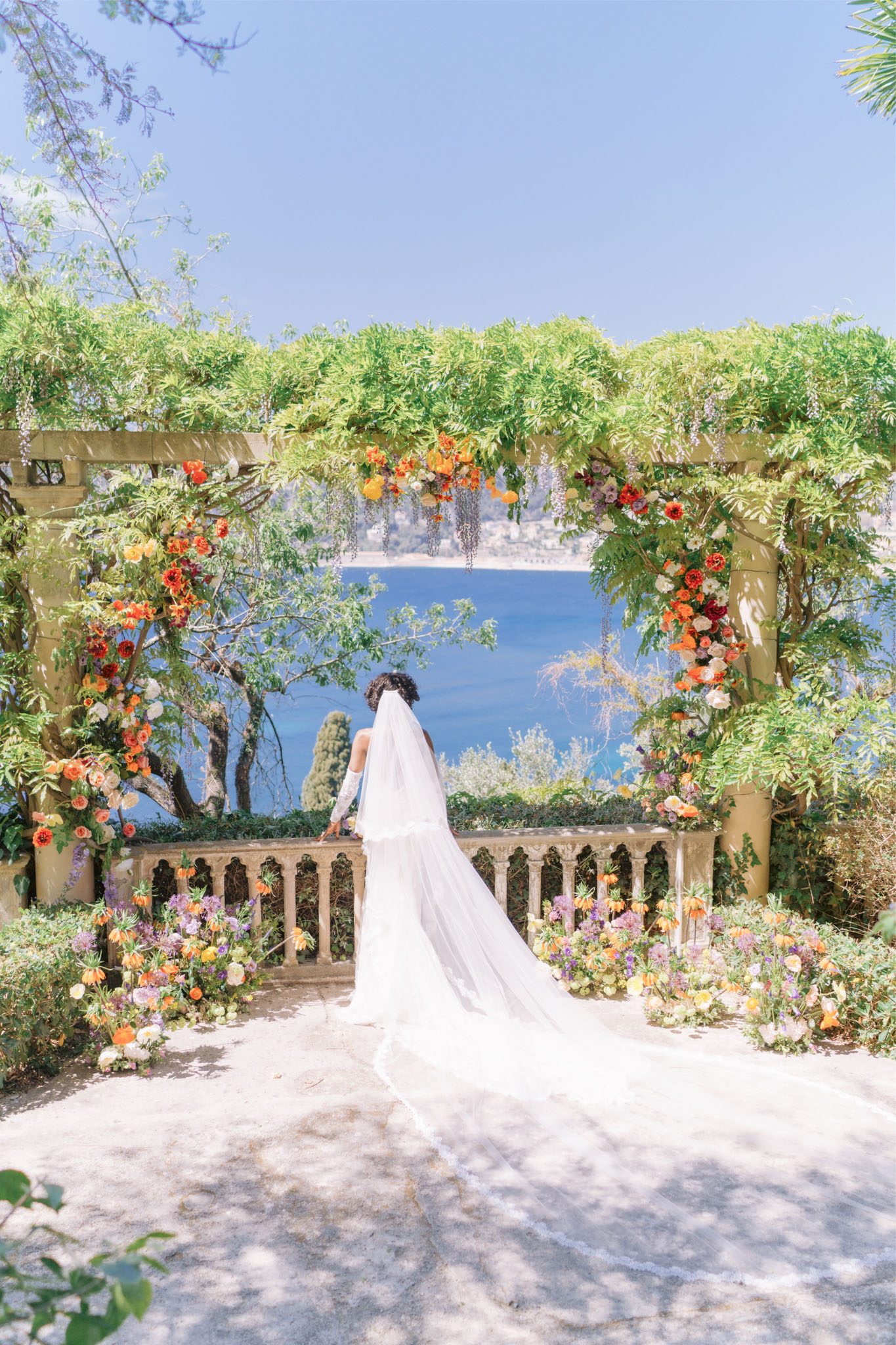 Bride in white gown and cathedral veil facing water view through vine-covered pergola with vibrant floral arch