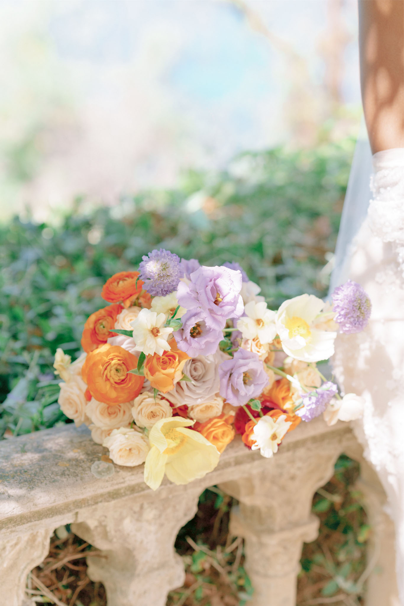 Bridal bouquet of orange ranunculus, lavender lisianthus, cream roses, and yellow tulips on stone bench