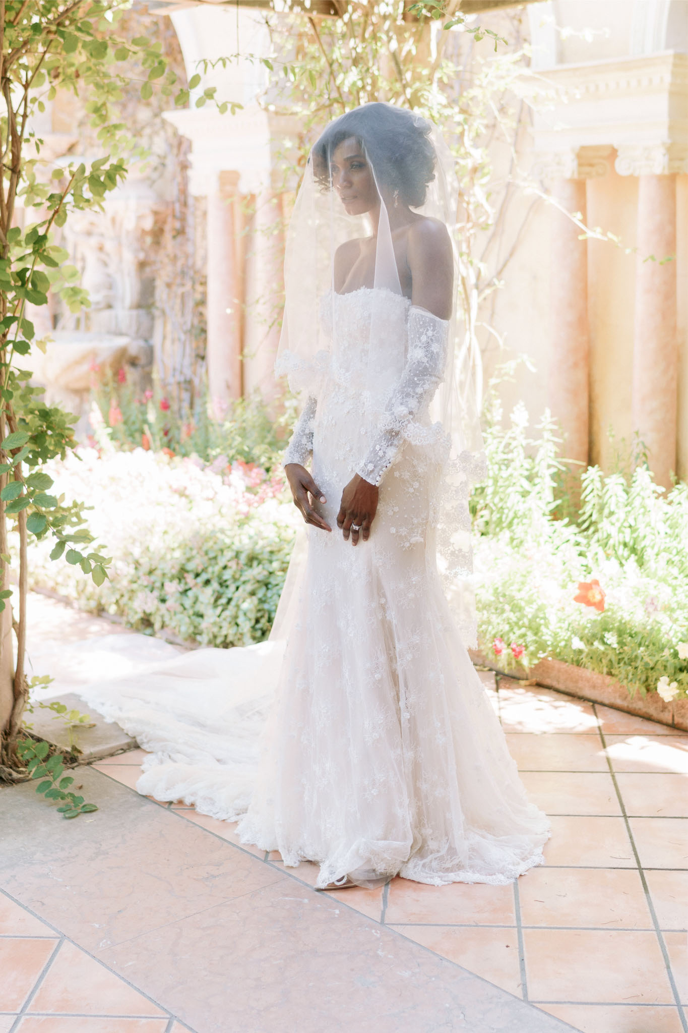 A bridal portrait of a single bride standing on a tiled terrace of what appears to be a classical-style venue with ornate stone columns and archways. She wears an ivory fitted lace gown with a mermaid silhouette, long lace sleeves, and an extended train, paired with a long cathedral-length veil draped over her upswept hair. She is turned slightly in profile, hands clasped at her waist, wearing a diamond engagement ring. The setting has a classic, formal architectural character with warm-toned stone columns visible in the background alongside blooming garden borders featuring red, pink, and white flowers. The image is a full-length portrait shot with bright, airy natural lighting.