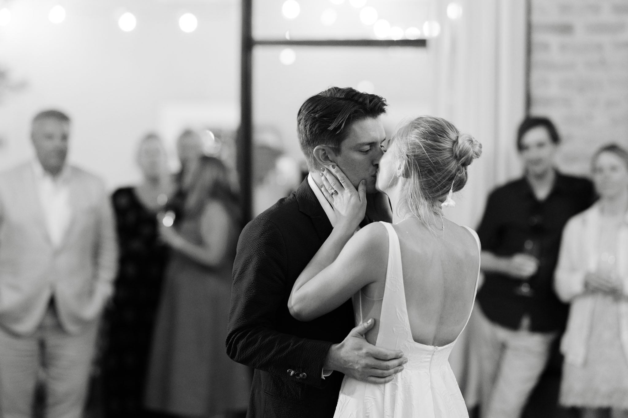 Black and white first dance kiss with string lights, bride in low-back gown holding groom's face