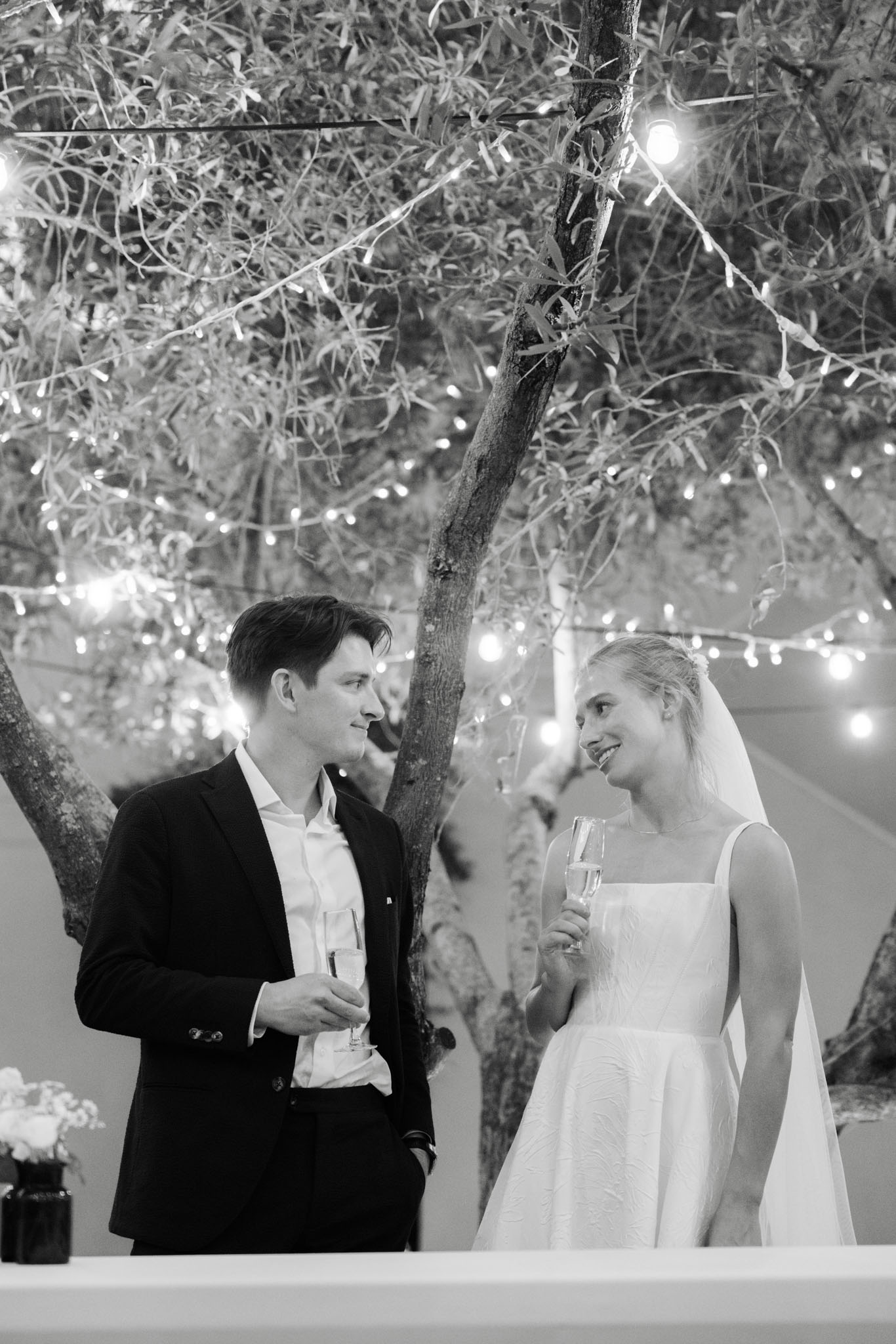 Black and white couple holding champagne flutes and smiling under fairy-lit tree at nighttime toast