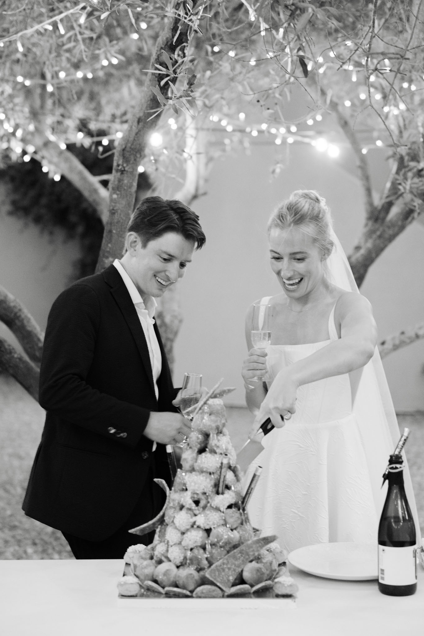 Black and white photo of couple cutting piece montee French choux tower under fairy lights at night