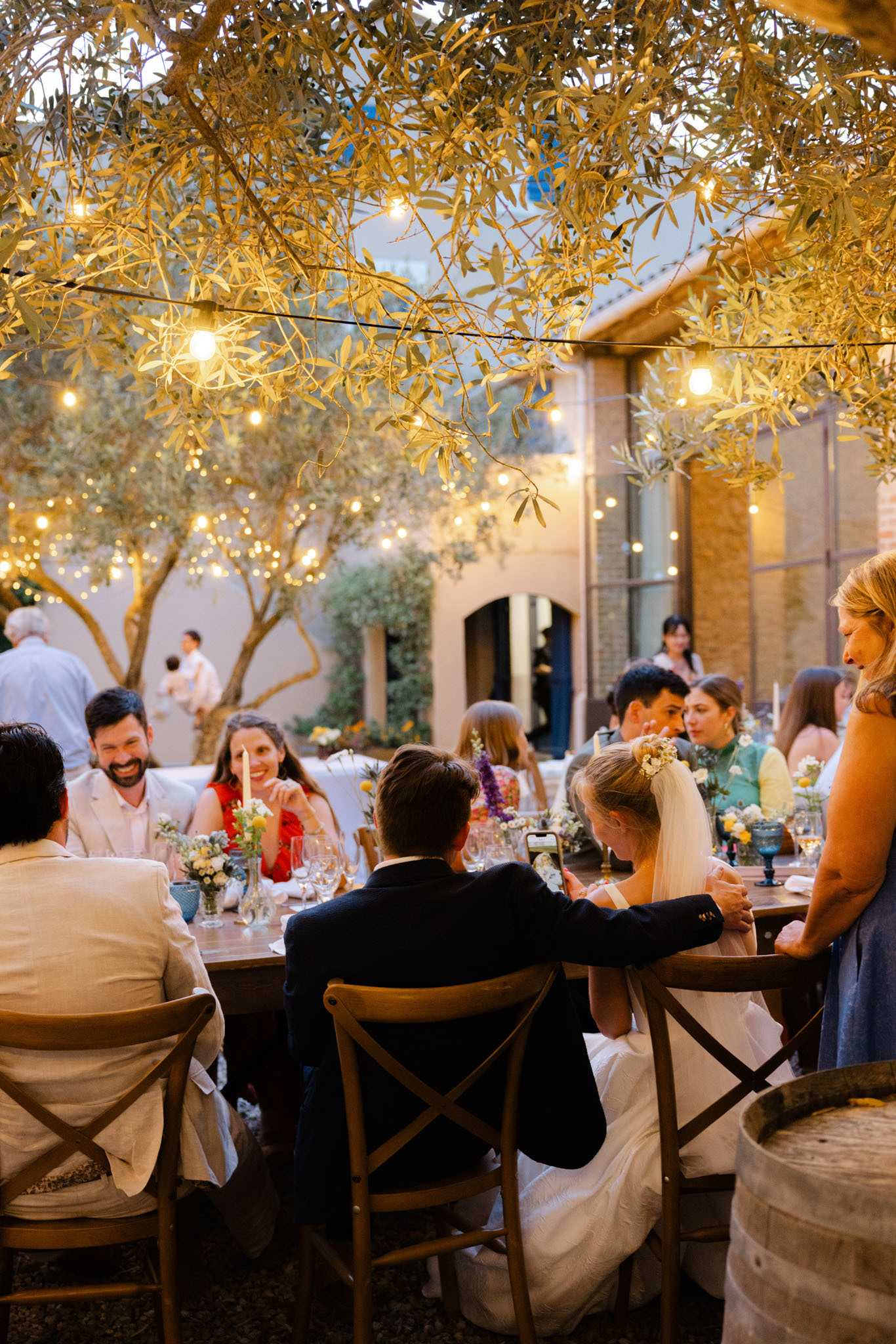 Evening reception under olive trees with string lights, wildflower bud vases, and taper candles at wooden table