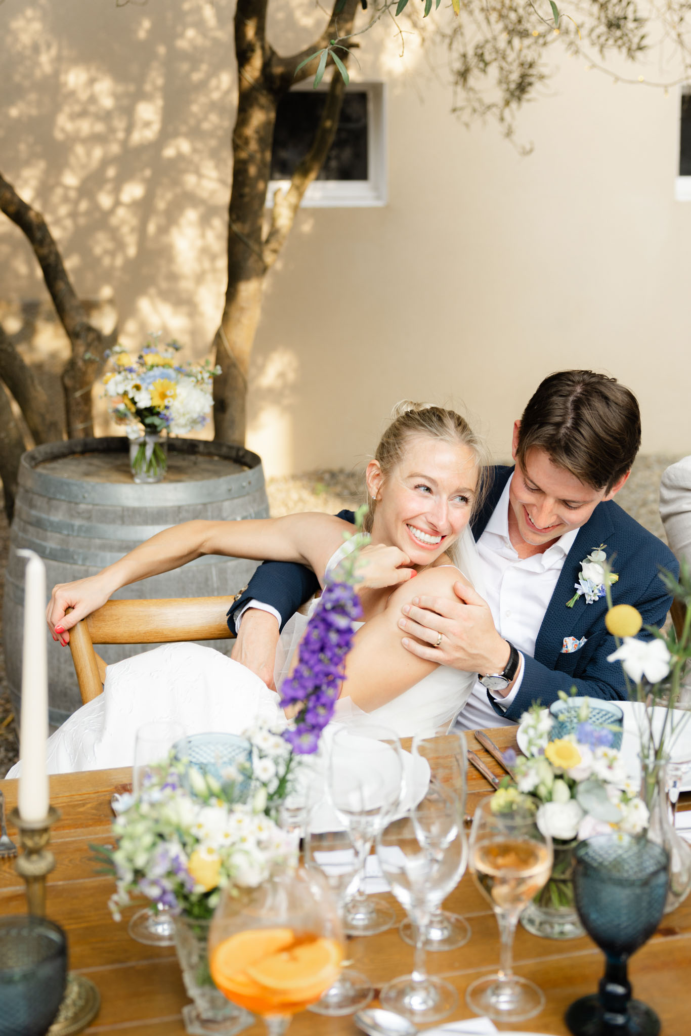 A couple sits together at their outdoor reception table, with the groom in a navy blue suit and white shirt with a boutonniere of white flowers, blue delphinium, and yellow craspedia leaning in to embrace the bride, who wears a white sleeveless dress and laughs toward the camera. The setting is an outdoor courtyard or terrace with a rendered cream wall and an olive tree in the background, and a wooden wine barrel holding a small floral arrangement of yellow, white, and blue wildflowers. The wooden dining table in the foreground is set with glass stemware including a glass of rosé wine, white taper candles in brass holders, blue textured goblets, and low floral centerpieces featuring white anemones, purple delphinium, yellow craspedia, and small white daisy-like blooms. The overall color palette is blue, yellow, and white with a relaxed Provençal style. The shot is taken from a slightly elevated angle, framing both the couple and the table decor.