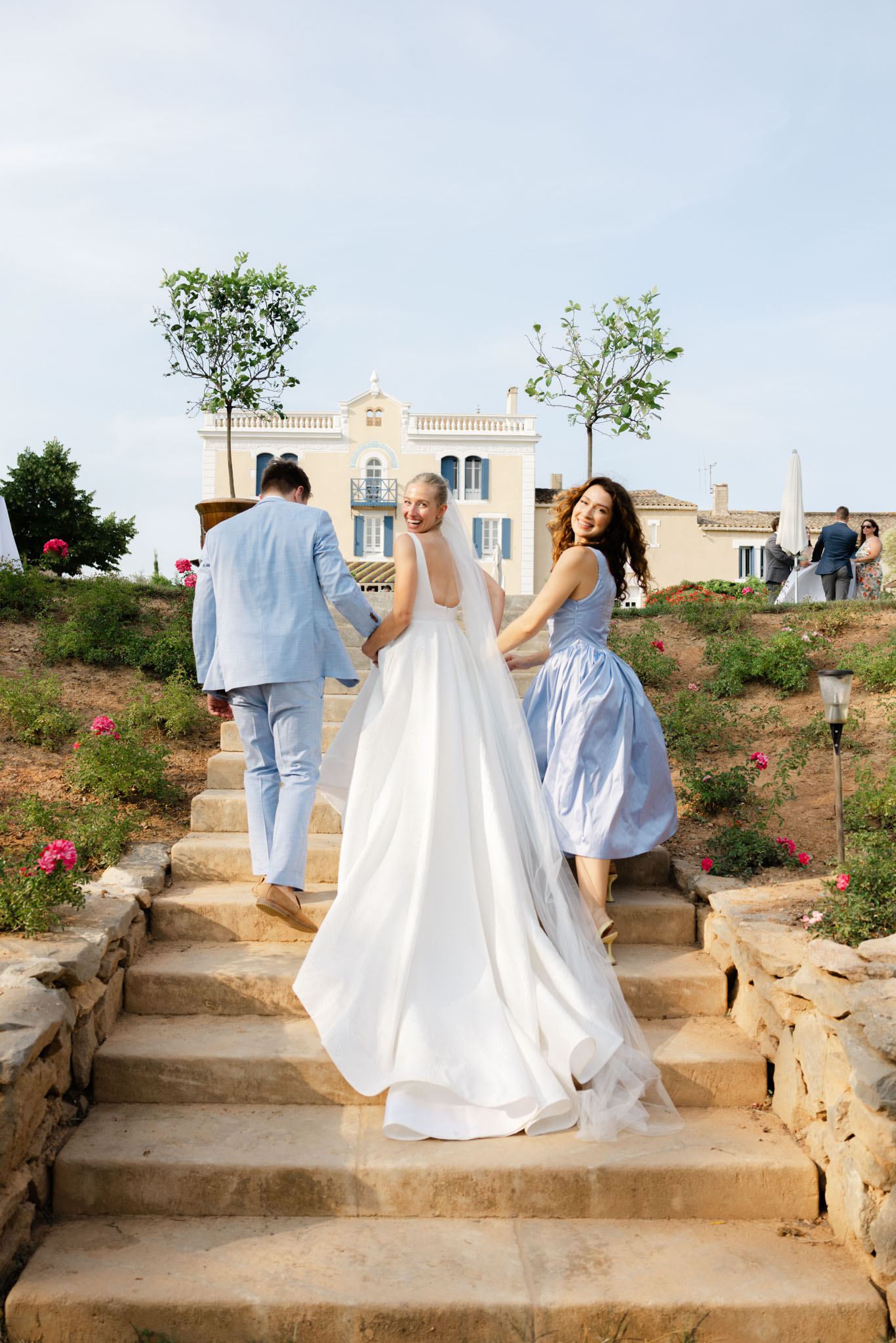 The bride, groom, and a bridesmaid walk up wide stone steps toward a yellow Provençal villa with blue shutters, likely during a transitional moment between the ceremony and reception. The bride wears a white full-skirted ballgown with an open back and a long veil, and turns to smile over her shoulder at the camera while the bridesmaid helps carry the train. The groom wears a light blue linen suit with tan shoes, and the bridesmaid wears a periwinkle blue midi dress with a fitted bodice. Several guests in smart-casual attire are visible in the background near the villa entrance, and pink flowering shrubs line the hillside steps. Wide shot, outdoor, classic French estate setting with a relaxed summer palette of white, light blue, and soft pink accents. Potential venue feature image.