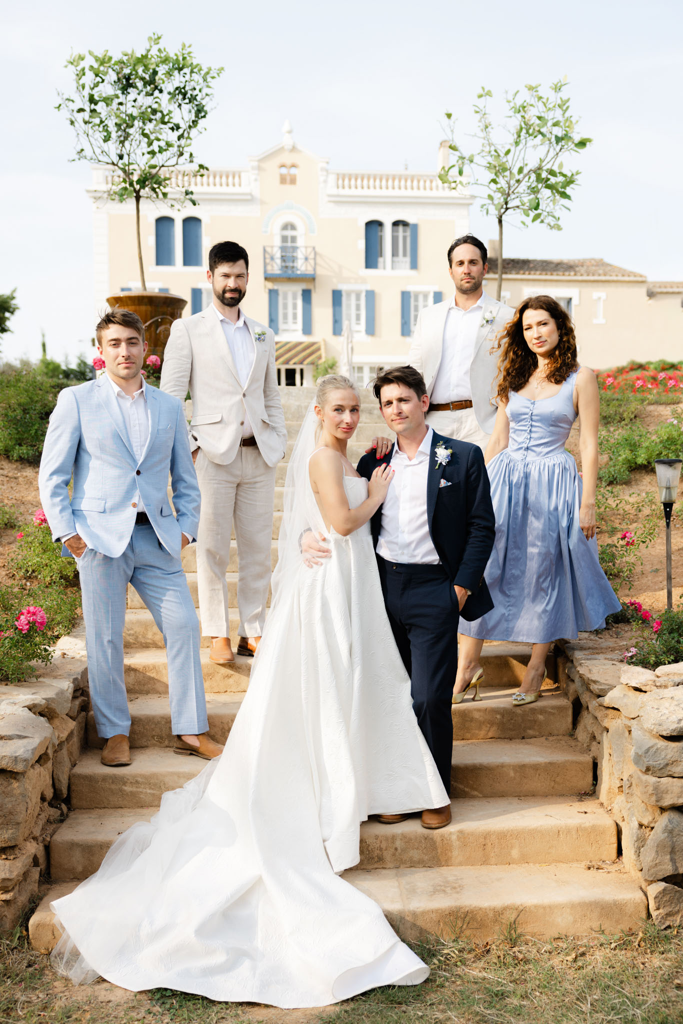 Bride, groom, and four bridal party members posing on stone steps of yellow Provencal villa with blue shutters