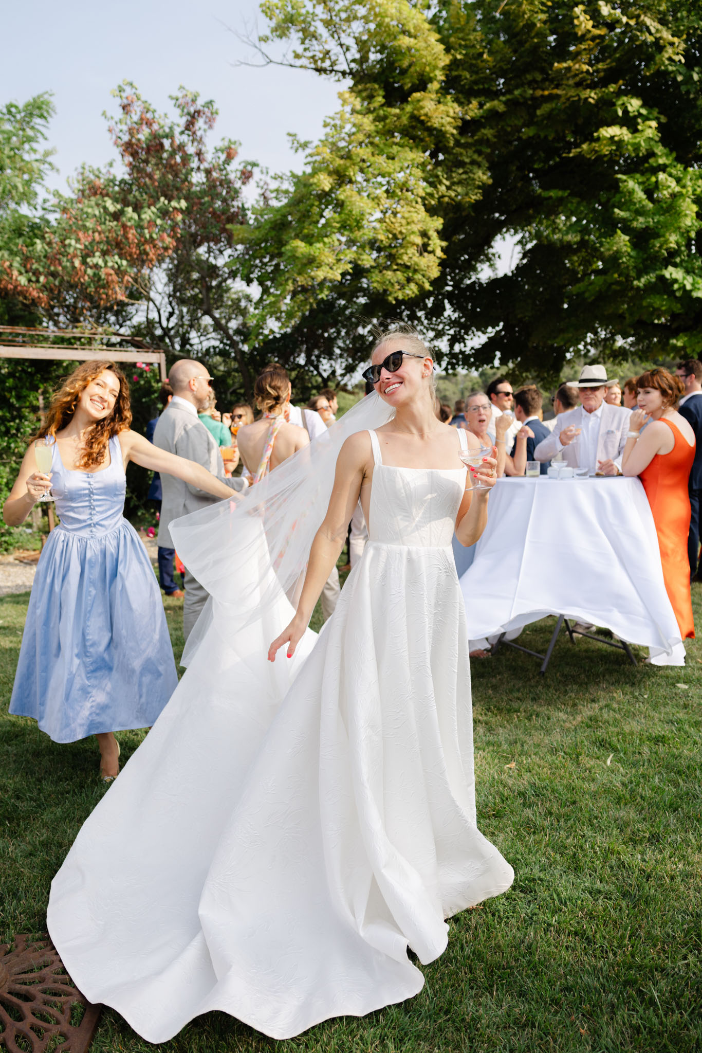 An outdoor cocktail hour scene on a lawn, with the bride in the foreground laughing and spinning, her white structured A-line gown with square neckline and spaghetti straps fanning out around her. She is wearing black cat-eye sunglasses, a long white veil, and holding a champagne coupe with red nail polish visible. Behind her, a woman in a periwinkle blue midi dress with a fitted bodice and full skirt is laughing and appears to be helping lift or toss the veil. Approximately 15–20 guests are visible in the background mingling around white cloth-covered cocktail tables, dressed in a mix of casual-formal attire including an orange dress, navy suits, and a white linen hat. The overall styling is modern and relaxed, with a candid, joyful energy captured in a medium-wide portrait shot.