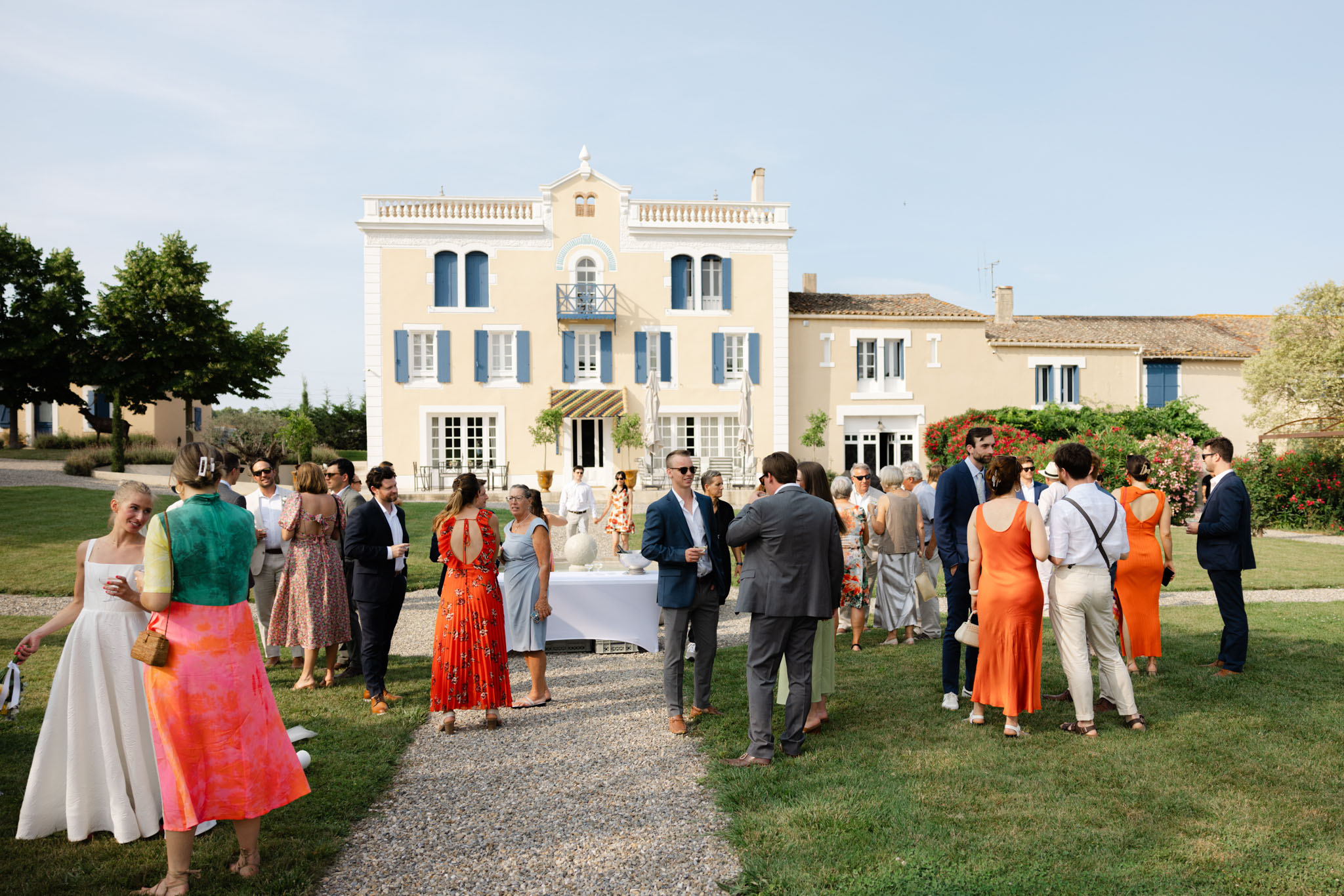 Wedding guests mingling on lawn during cocktail hour in front of yellow French manor house with blue shutters