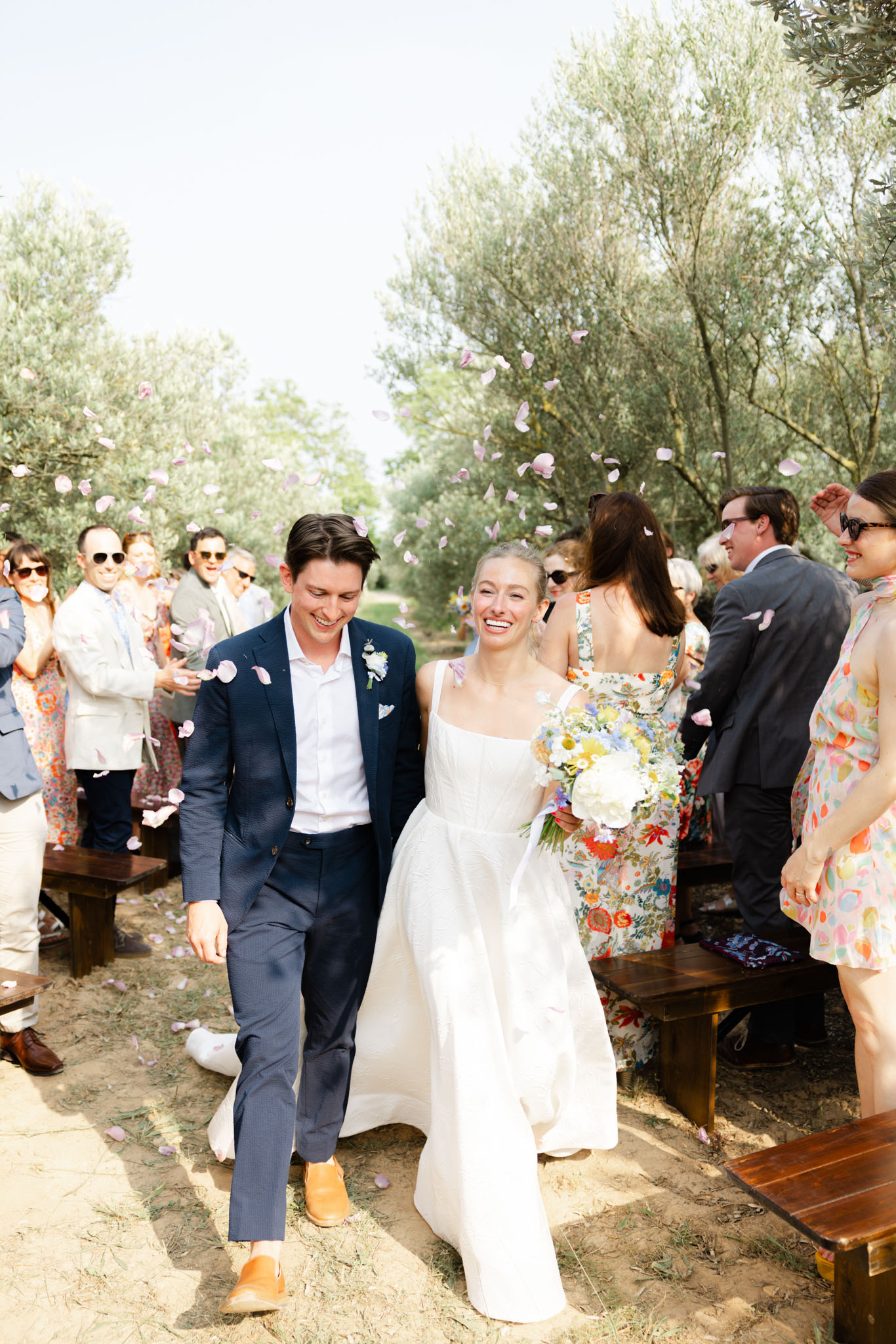 Couple walks up aisle as guests throw pink petals during outdoor ceremony recessional lined with olive trees