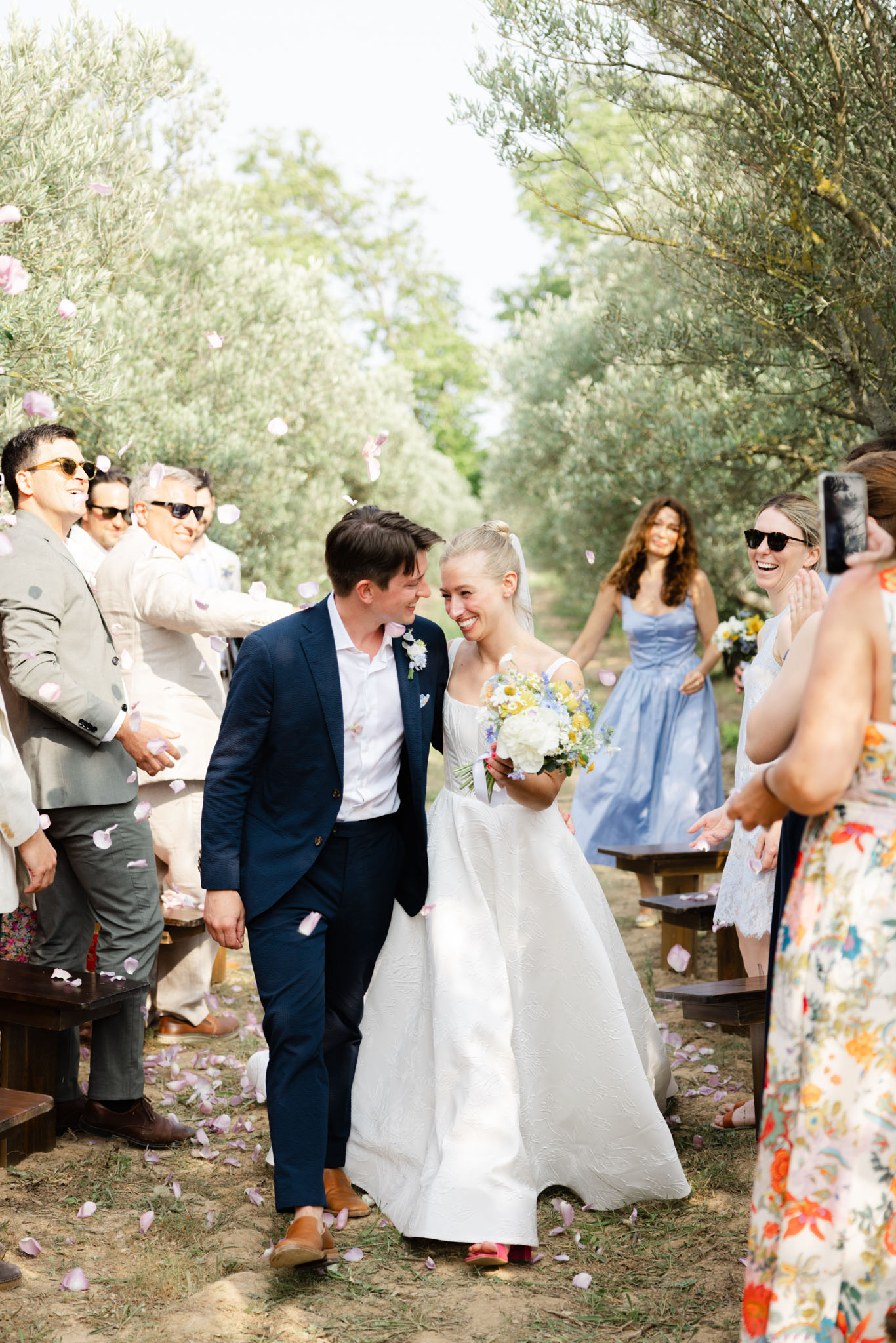 The couple walks back down the aisle together following an outdoor ceremony held in an olive grove, with guests lining either side tossing pink flower petals. The groom wears a navy suit with a white shirt and tan leather shoes, with a small white boutonniere on his lapel, while the bride wears a white structured ball gown with a square neckline and carries a mixed bouquet of white peonies, yellow chamomile, blue daisies, and greenery. Approximately 10–12 guests are visible, dressed in a variety of casual-smart attire including a periwinkle blue midi dress, a floral print dress, and linen separates; most are smiling and laughing, with one guest filming on a phone. The ceremony seating consists of dark wooden benches placed along the dirt aisle, and pink petals are scattered across the ground, giving the setting an informal, relaxed feel. Medium-wide portrait shot captured in natural daylight.